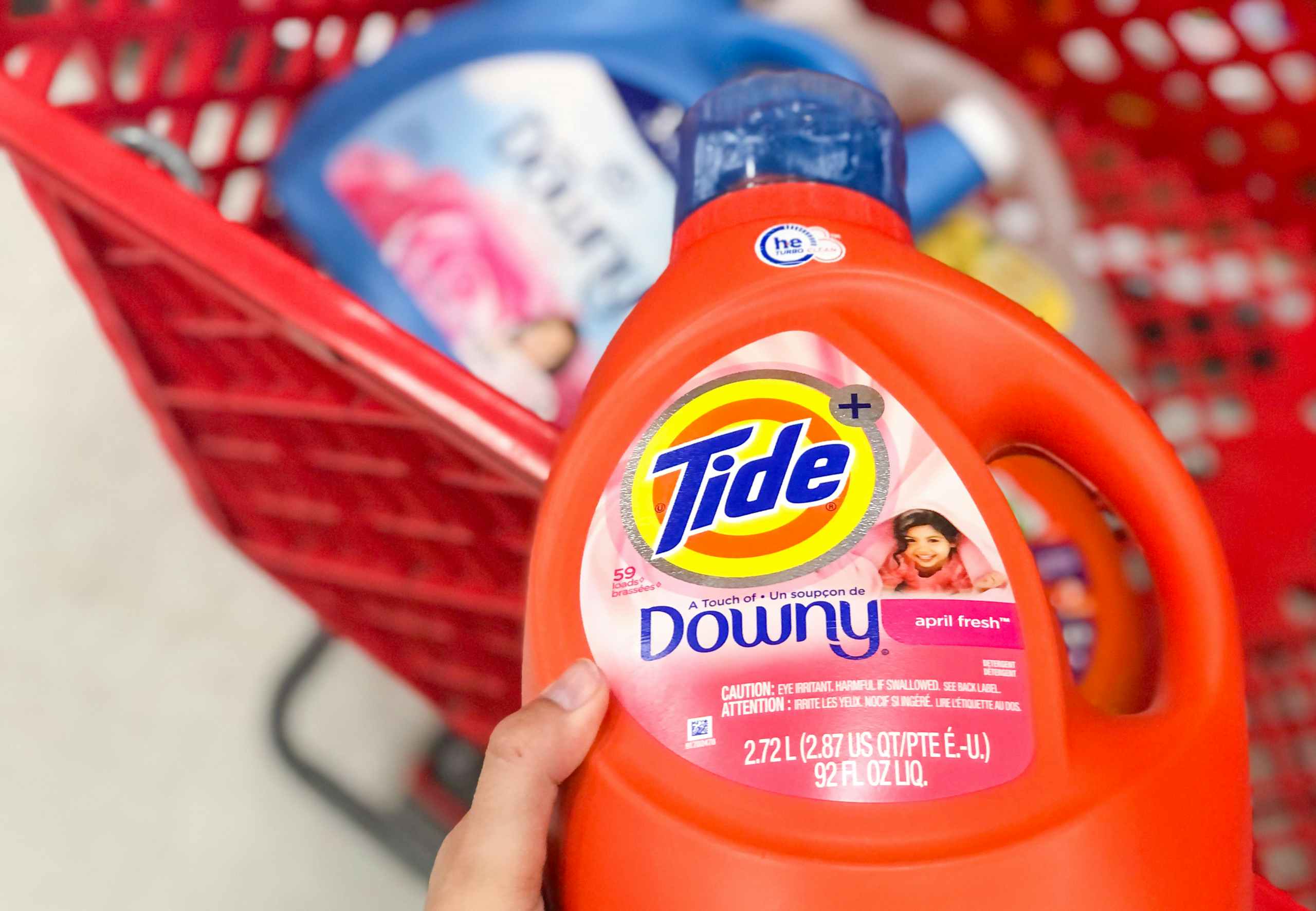 hand holding bottle o tide detergent over shopping cart with other bottles of detergent