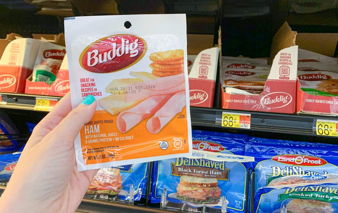 hand holding packet of buddig lunch meat in front of sliced meats in walmart fridge area
