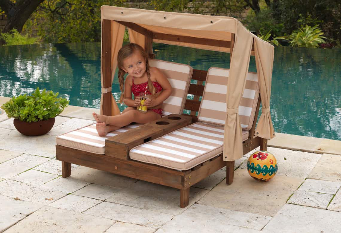 stock photo of little girl sitting on kidkraft double chaise lounge chair with cupholders and shade in front of shaded pool