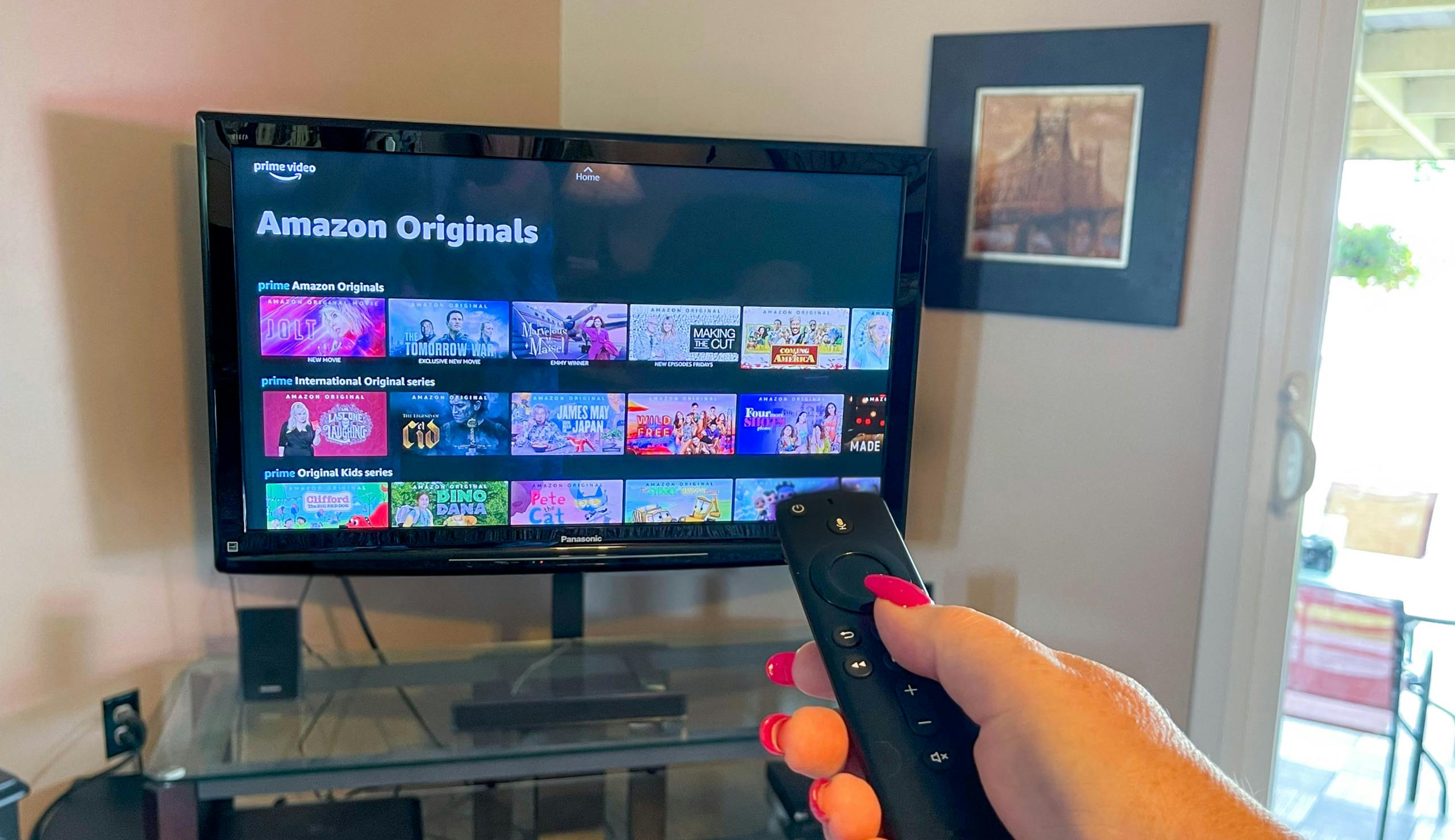 A person's hand holding an Amazon Fire remote, aiming it at a TV displaying Prime Video's Amazon Originals. 