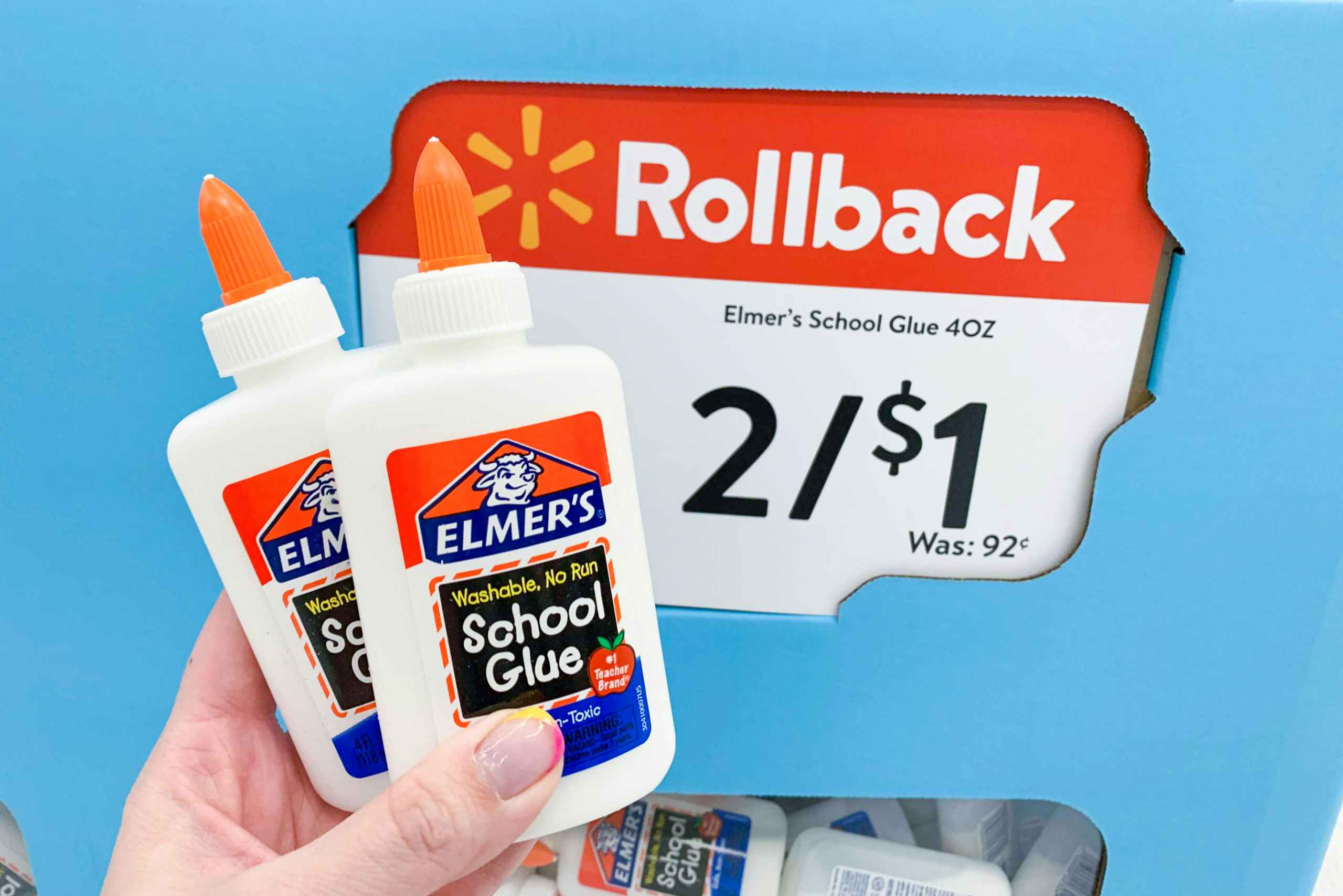 A person's hand holding 2 bottles of Elmer's School Glue in front of a Walmart Rollback sale sign that reads, "Elmer's school glue 4oz, 2/$1" inside Walmart.