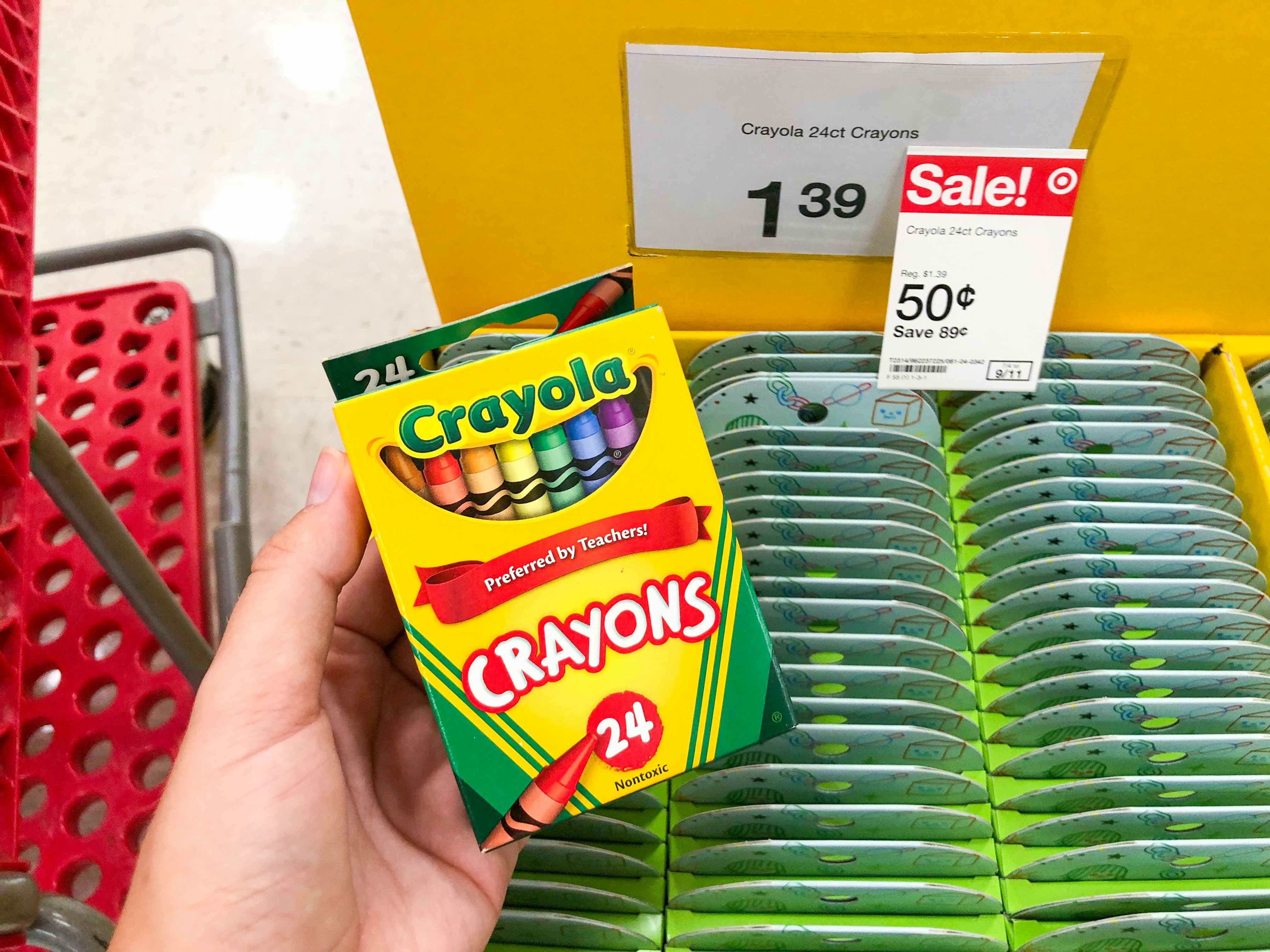 A person's hand holding a box of Crayola crayons in front of a sale tag marking them down to 50 cents each at Target.