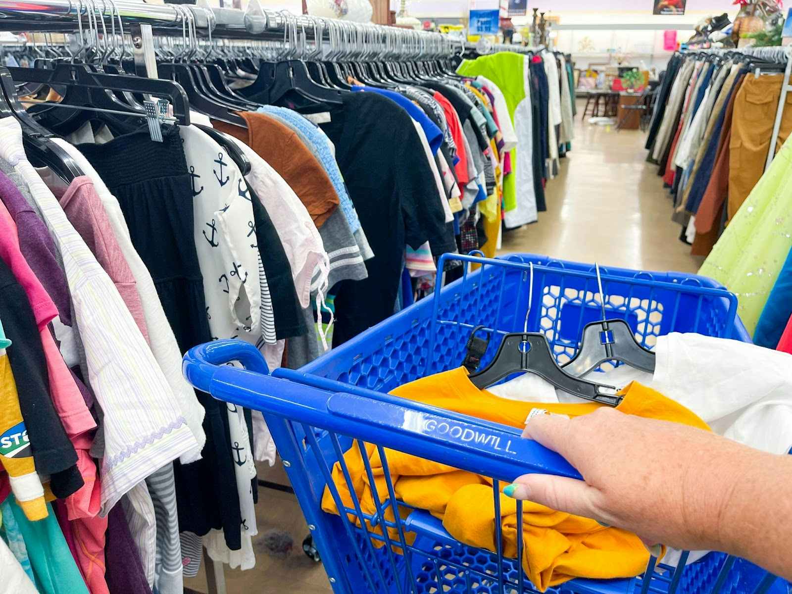 A person with their hand on a shopping cart in a clothing aisle of a goodwill store.