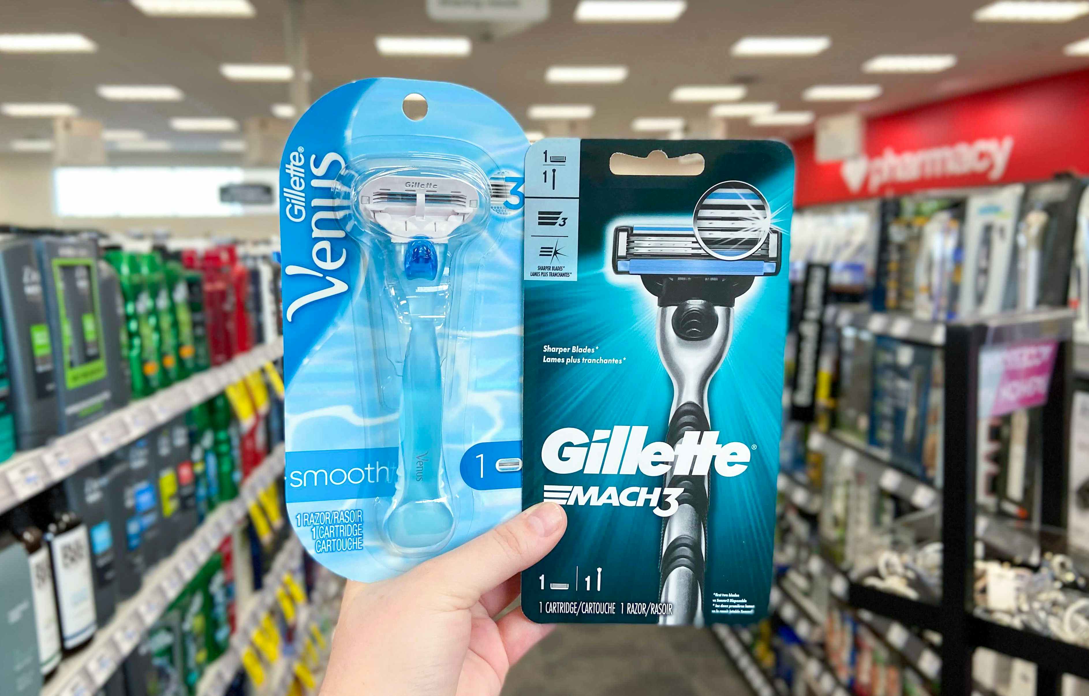 A person's hand holding up two reusable Gillette razors in front of an aisle at a store. One is a Venus women's smooth razor and the other is a Mach 3 men's razor.