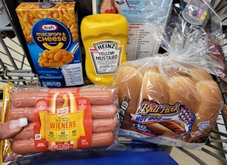 A box of Kraft mac and cheese, a package of Oscar Mayer wieners, a bottle of Heinz yellow mustard, and a bag of Ball Park hotdog buns in the basket of a shopping cart. 