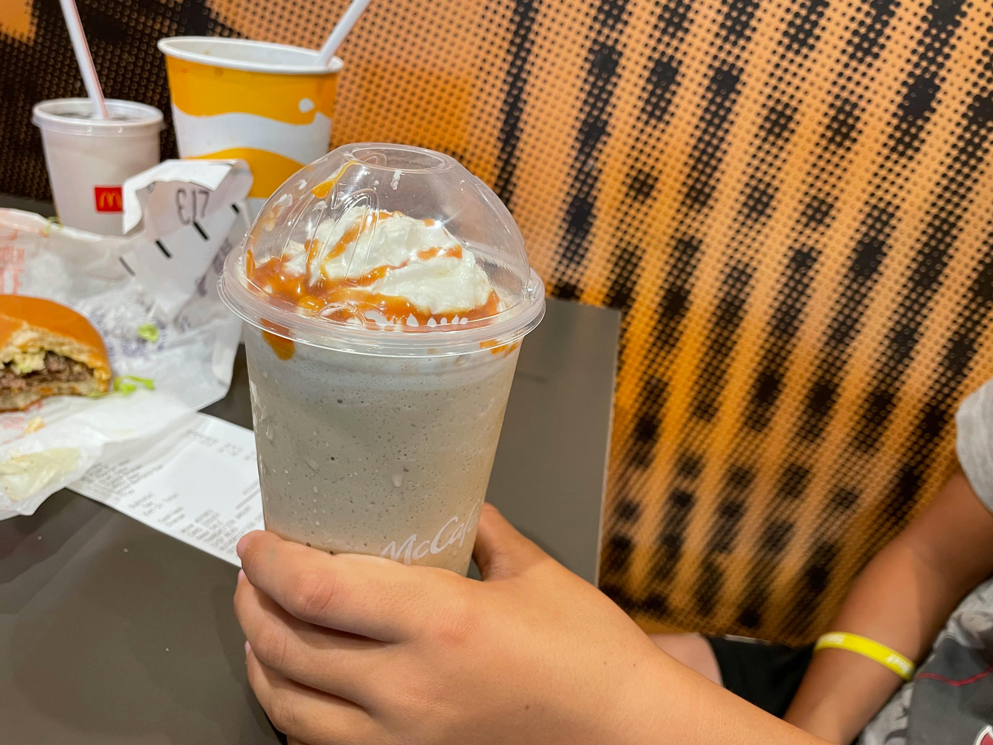 A person holding a McDonald's large caramel frappe while sitting at a table inside McDonald's.
