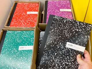A person's hand taking a composition notebook out of a box of composition notebooks at Office Depot.