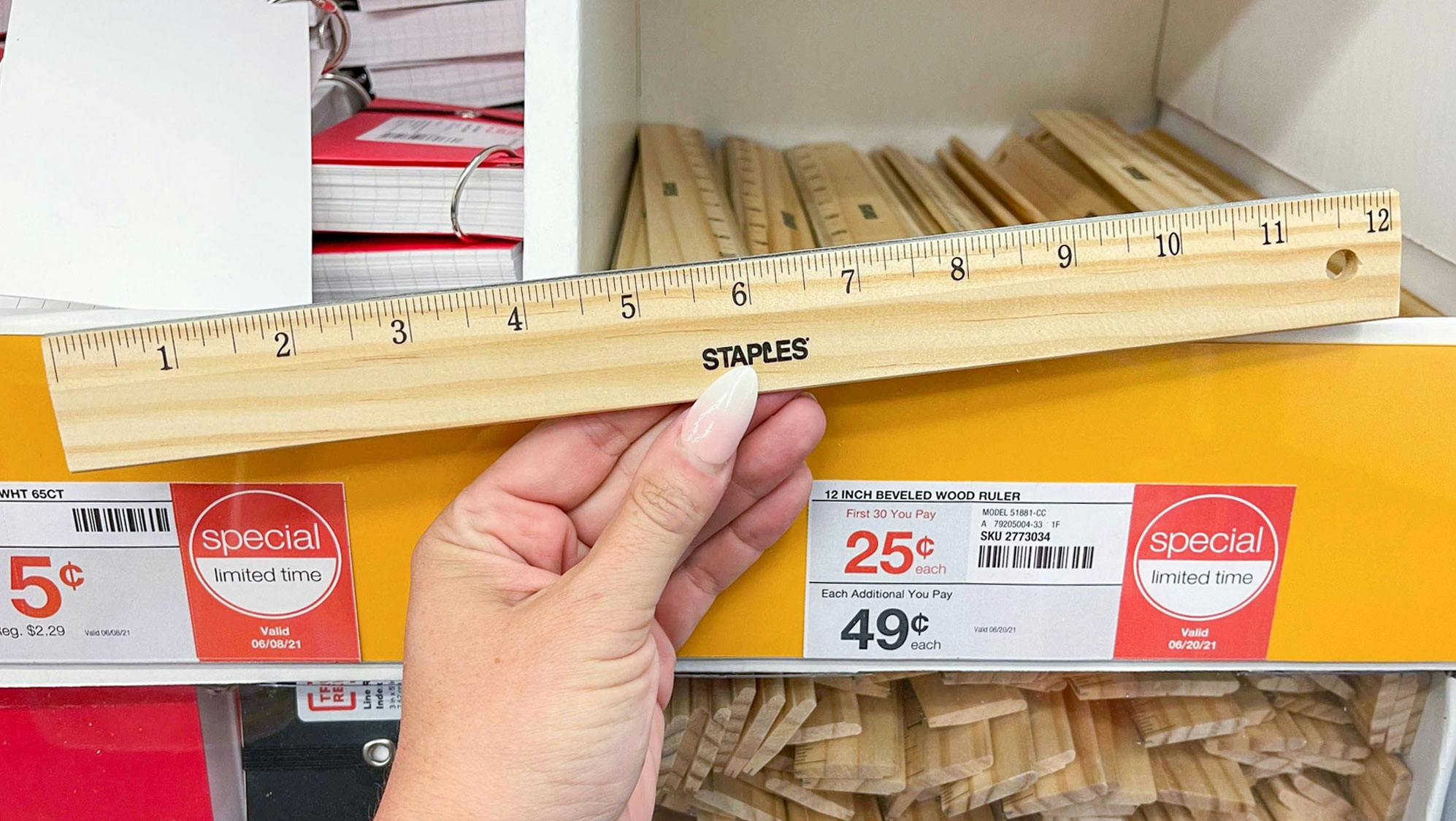 A person's hand holding a Staples ruler in front of a shelf with more rulers on sale for $0.25 at Staples.