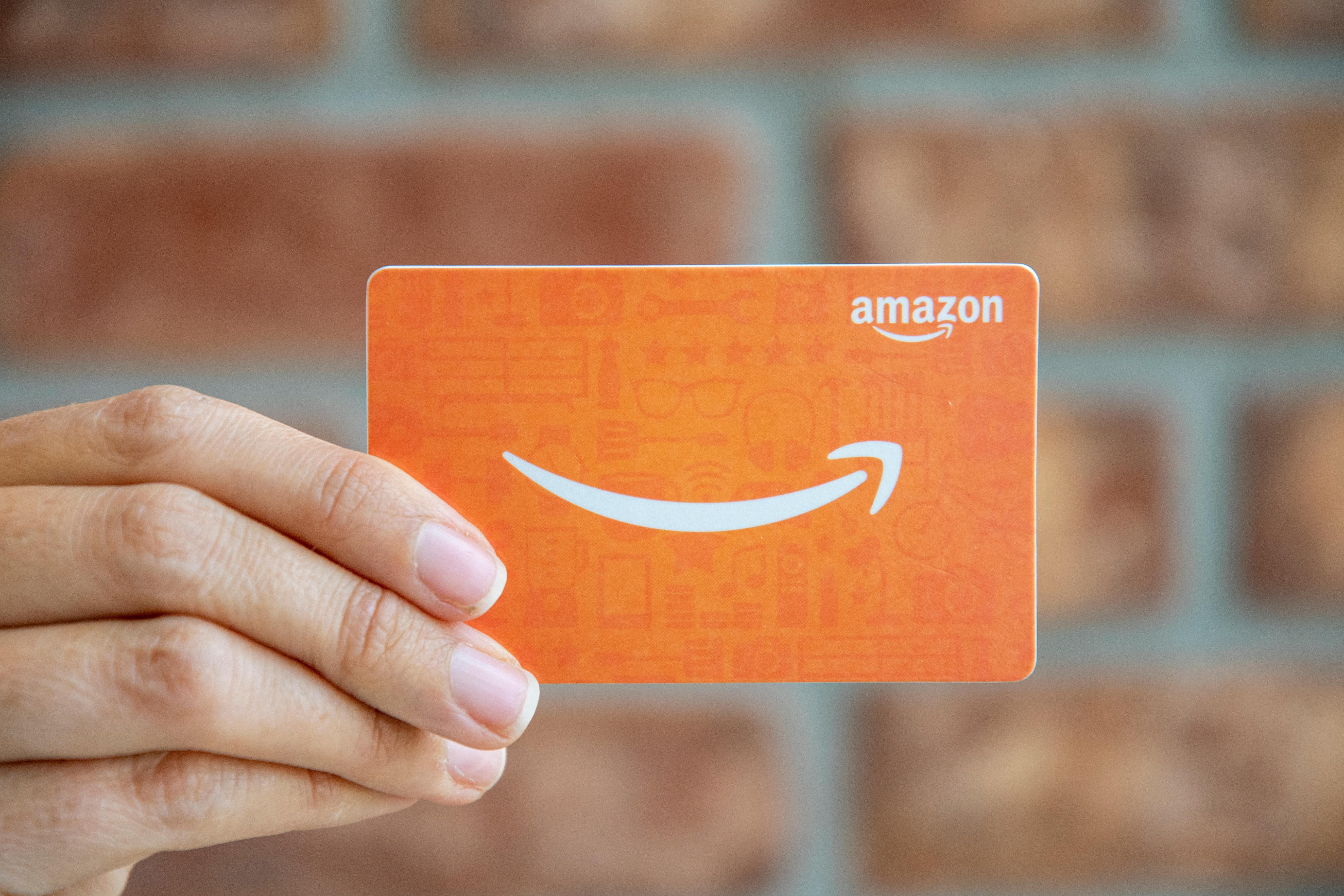 A person's hand holding up an Amazon gift card in front of a brick wall.