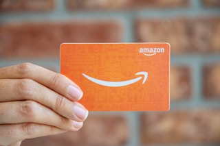 A person's hand holding up an Amazon gift card in front of a brick wall.
