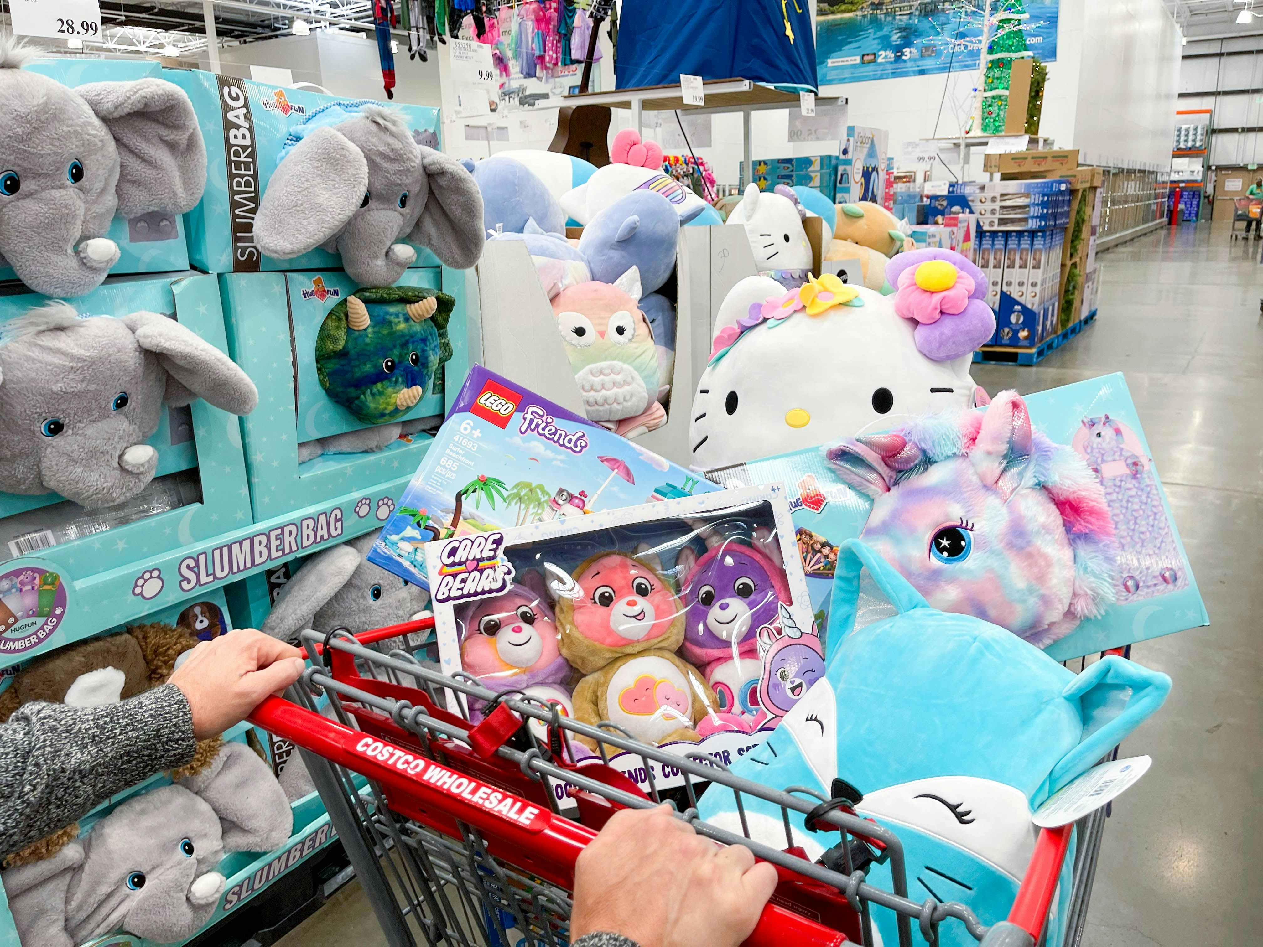 A person pushing a Costco shopping cart with the basket filled with plush toys in the toy aisle at Costco.