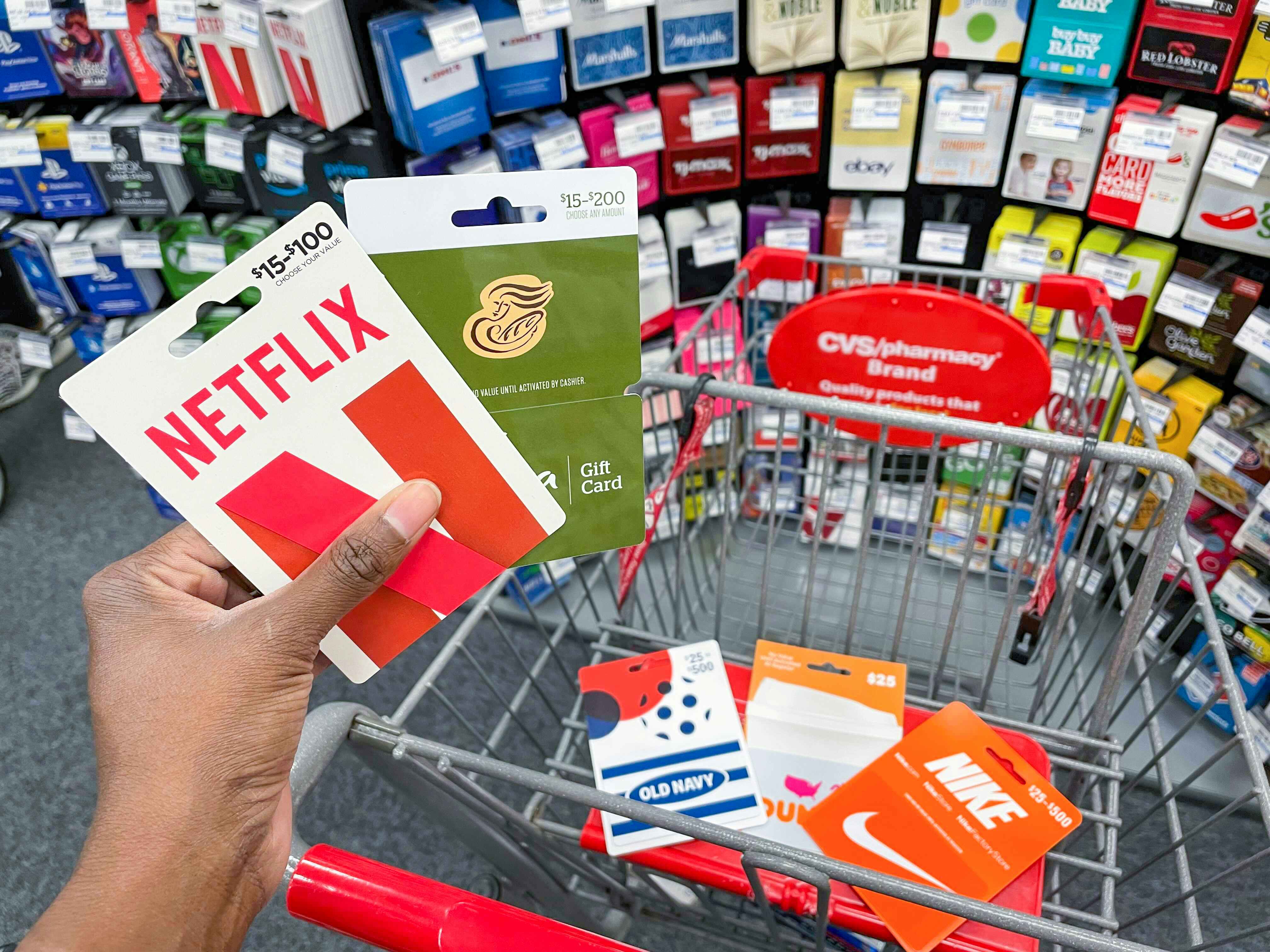 A person's hand holding a Netflix and a Panera gift card next to a CVS shopping cart with more gift cards in the basket, parked in front of the gift card display inside CVS.