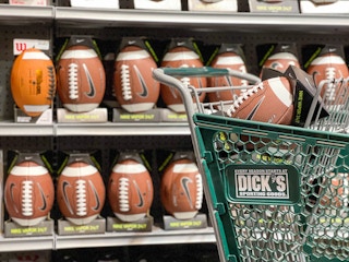 Footballs on a for sale shelf with one in a Dicks sporting goods shopping cart in the forground