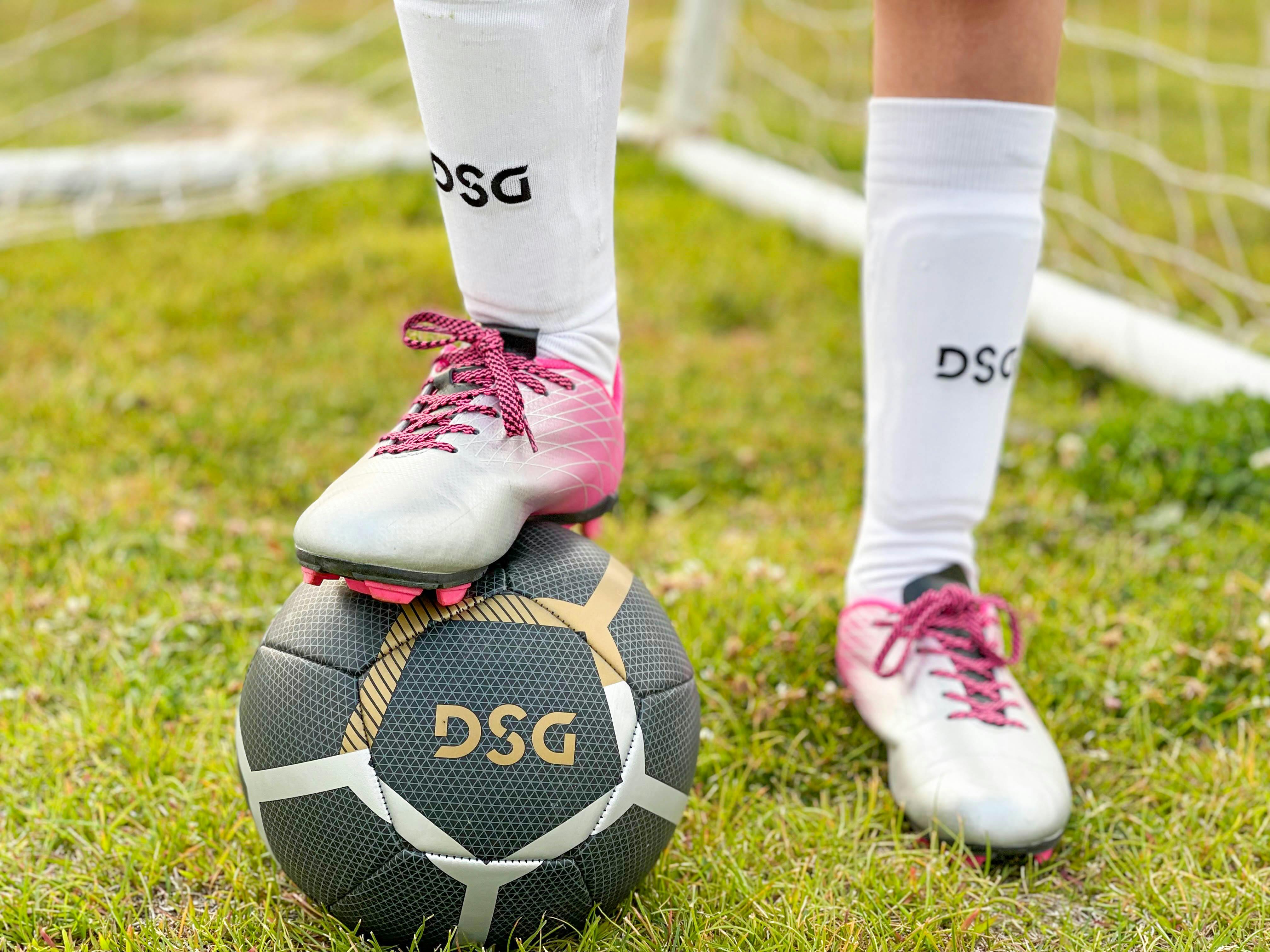 A child wearing DSG soccer shoes, socks, pads, stepping on a DSG soccer ball.