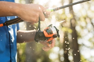 A person holding a fishing rod with water spraying up around them and sun coming through the trees in the background