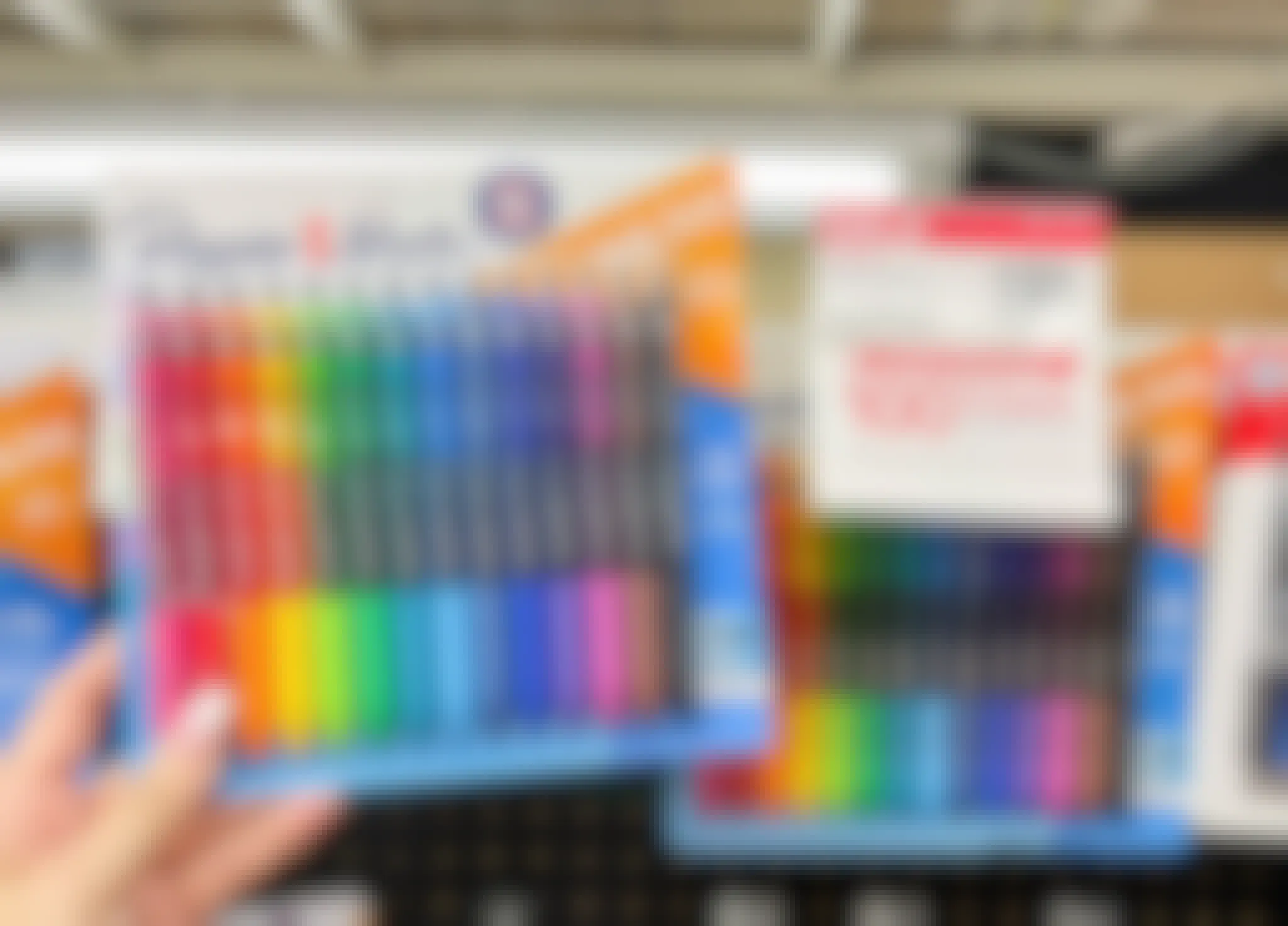 A person's hand holding a 14-pack of PaperMate gel pens in front of a display with more pens at Office Depot.