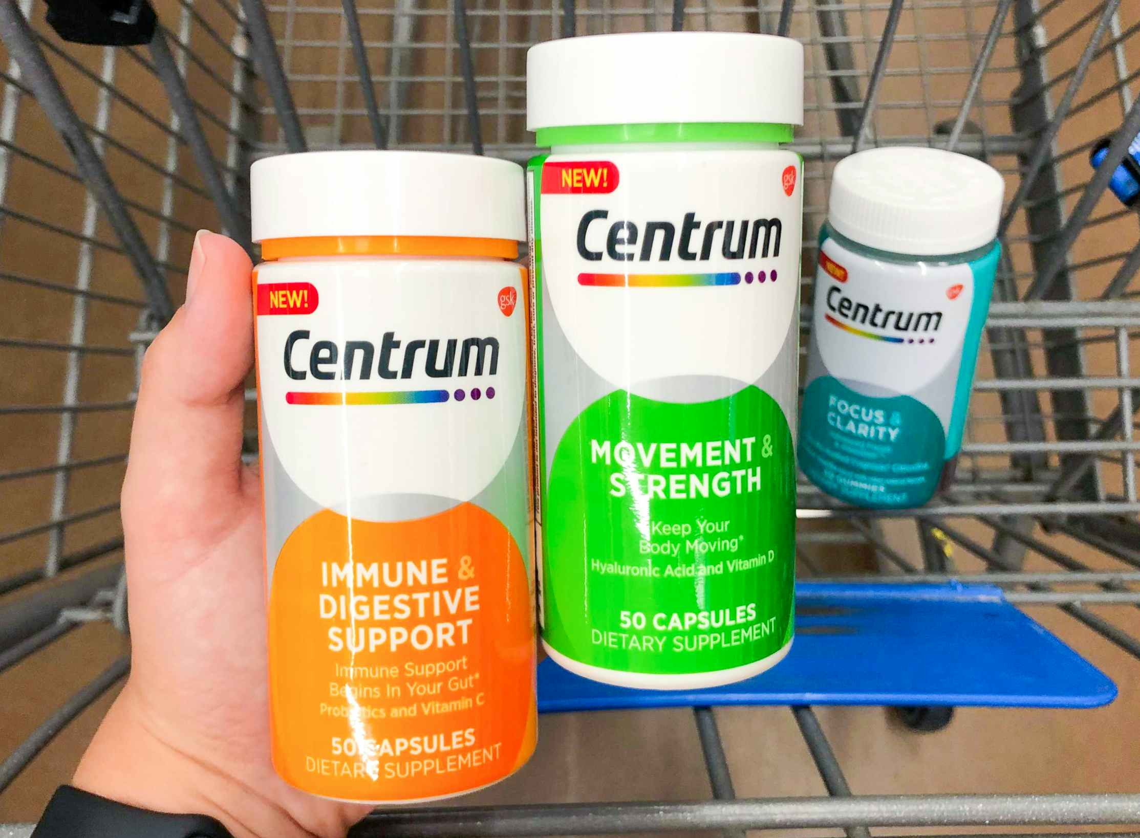 A person's hand holding two bottles of Centrum vitamins over a shopping cart basket with another bottle sitting in it.