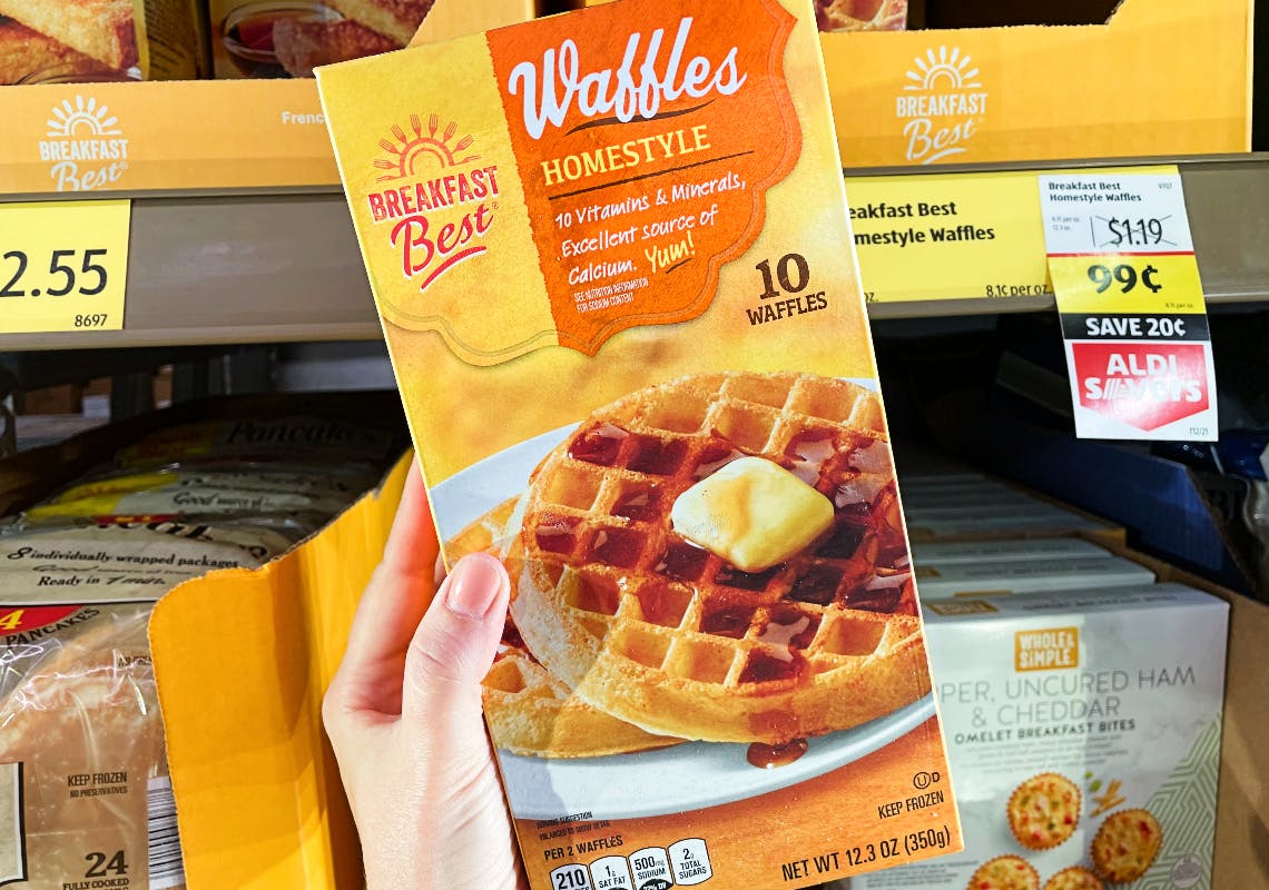 Hand holding a frozen waffles box in front of the freezer section of a grocery store.