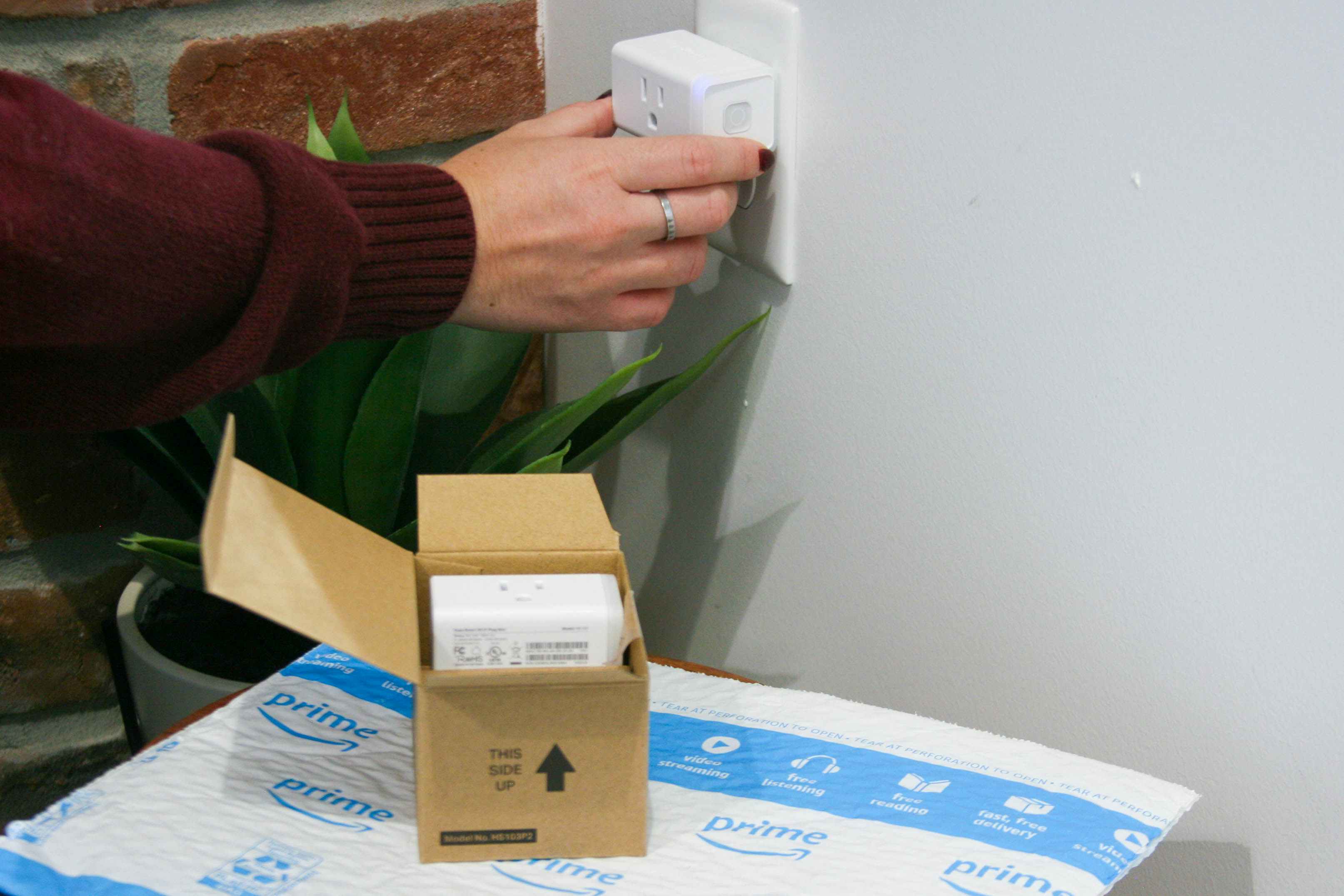 A person plugging a smart plug into an outlet next to a box of more smart plugs on top of an Amazon shipping envelope.