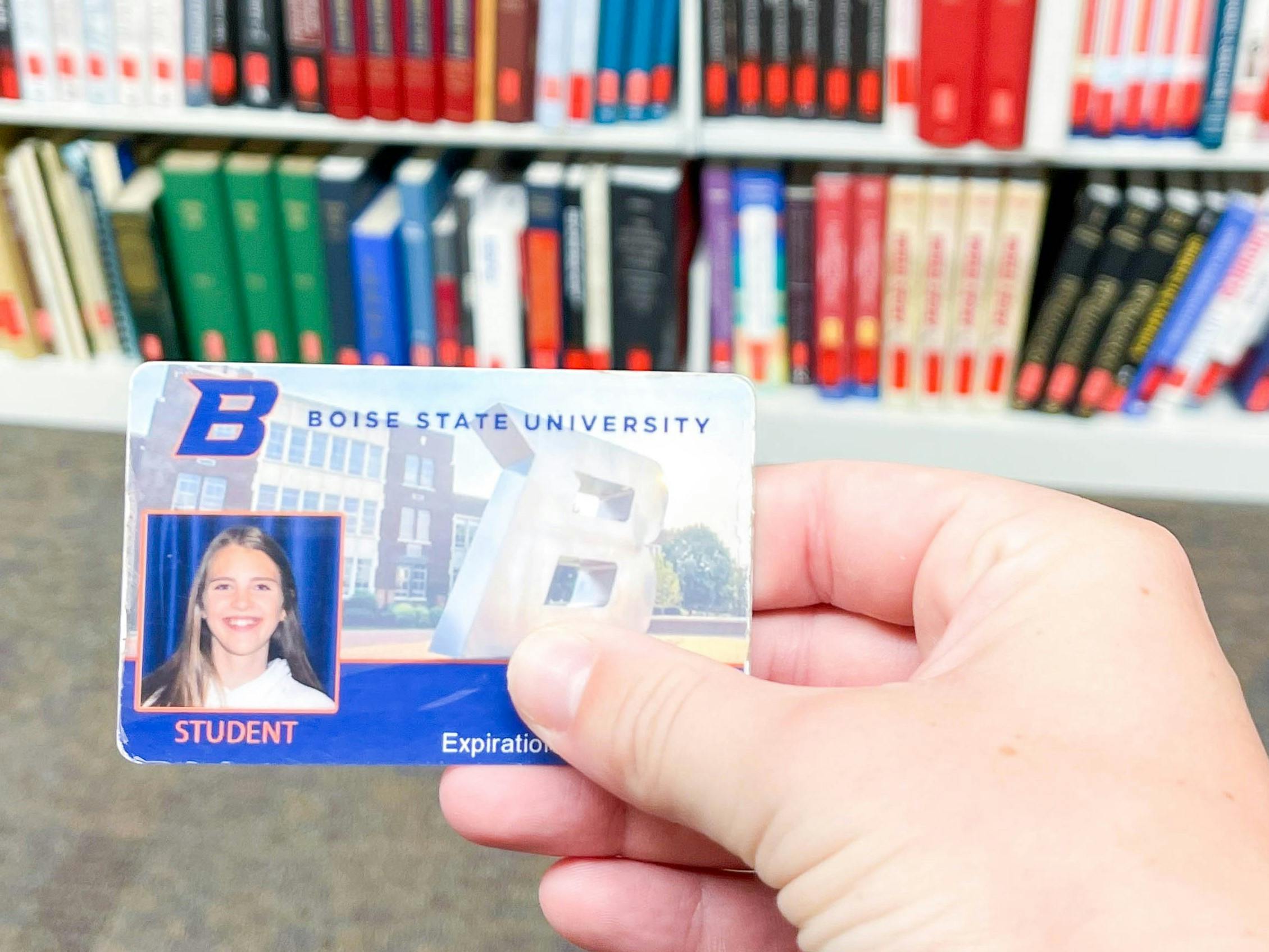A college student holding their student ID in front of a bookshelf in a library.