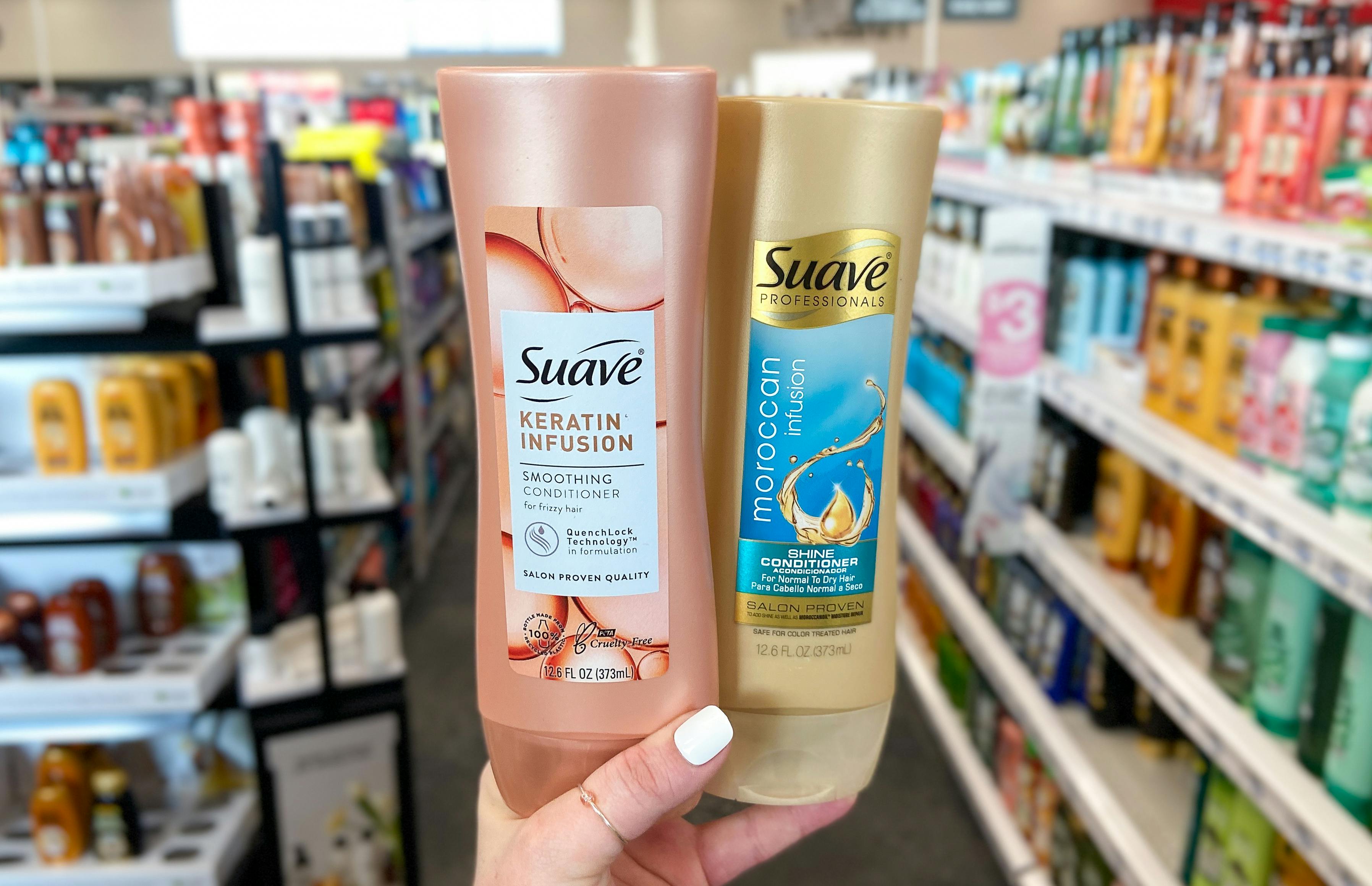 A person's hand holding up two bottles of Suave conditioner in front of an aisle in a store. One is Suave Keratin Infusion smoothing conditioner and the other is Suave Moroccan Infusion shine conditioner.