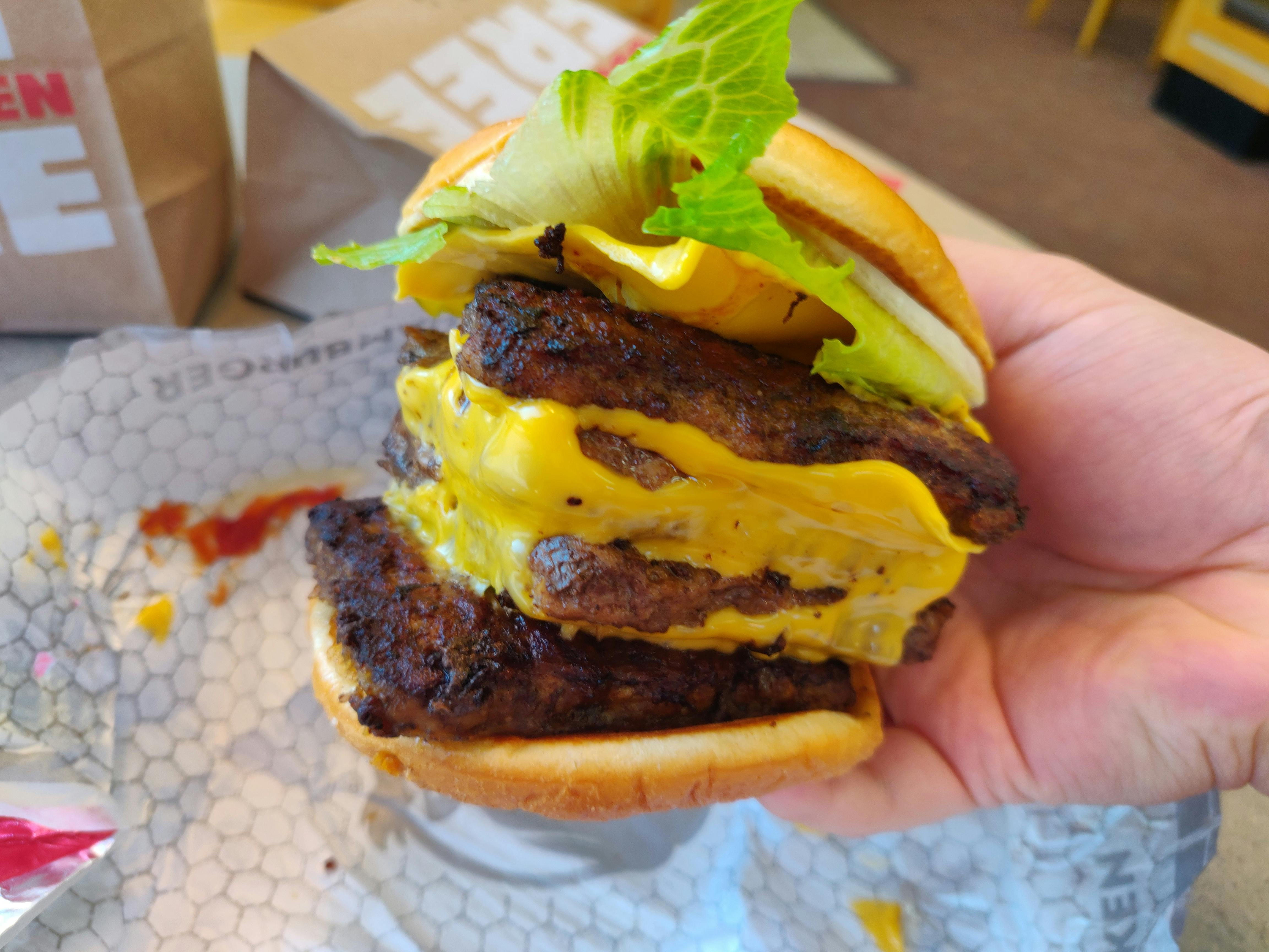 A person's hand holding up a Wendy's Meat Cube (four patties stacked on top of each other) above a paper wrapper on the table at Wendy's.