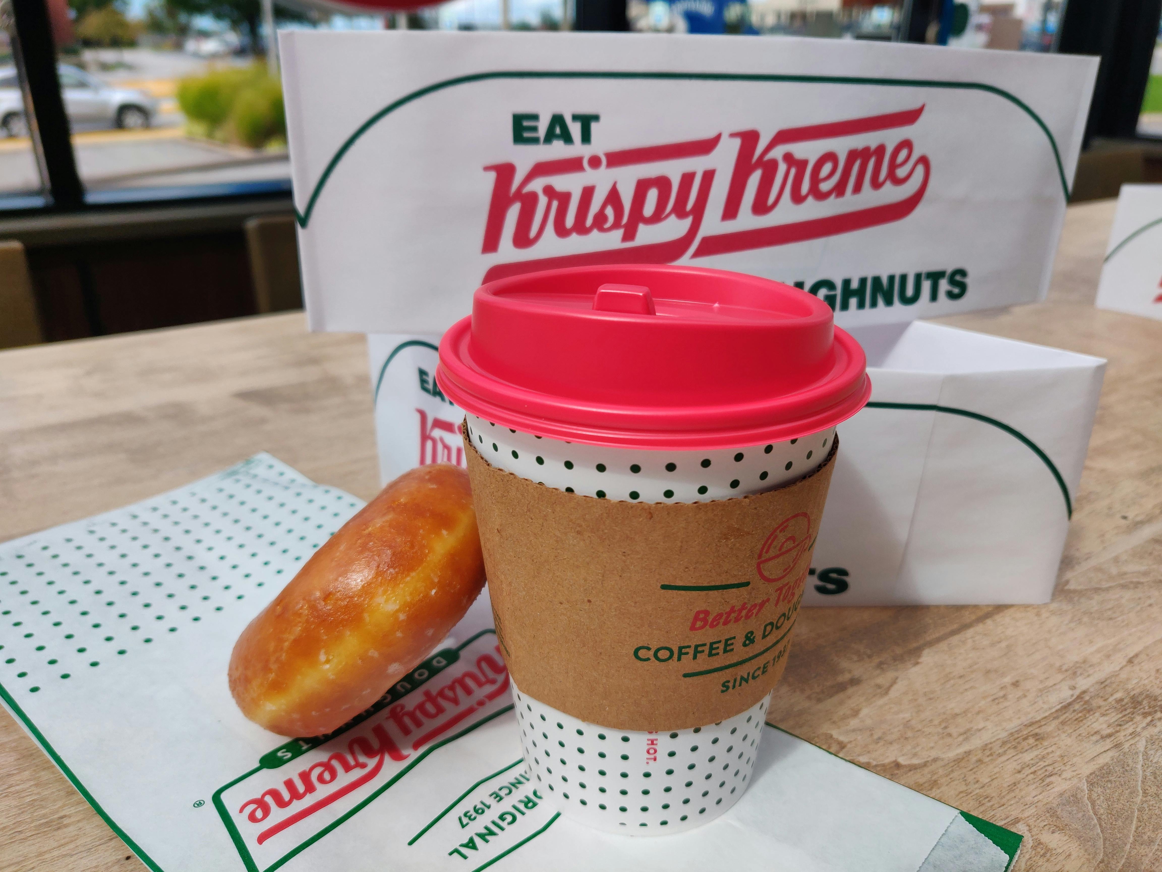 A Krispy Kreme doughnut sitting on top of a Krispy Kreme bag with a small coffee and Krispy Kreme hats in the background..