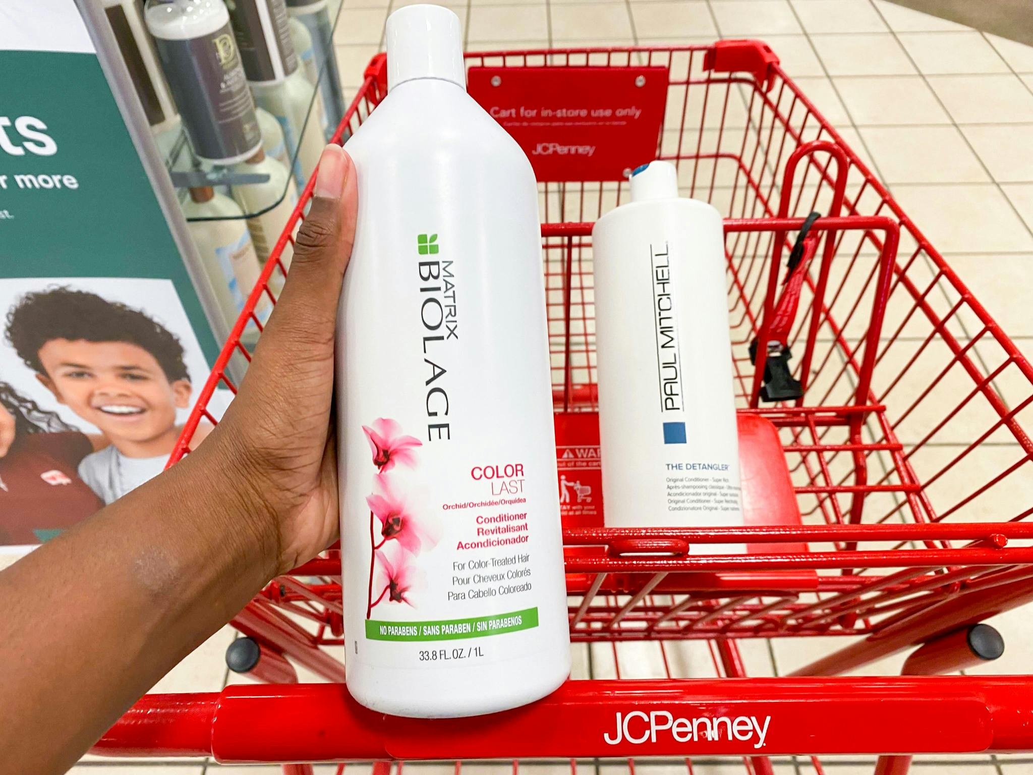 A person's hand holding a bottle of Matrix Biolage shampoo in front of a bottle of Paul Mitchell shampoo sitting in a JCPenney shopping cart basket.