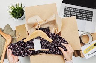 A person's hands taking a dress on a hanger out of a cardboard box that is sitting on a desk next to a laptop and a plant.