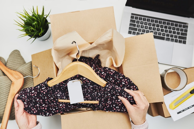A person's hands taking a dress on a hanger out of a cardboard box that is sitting on a desk next to a laptop and a plant.