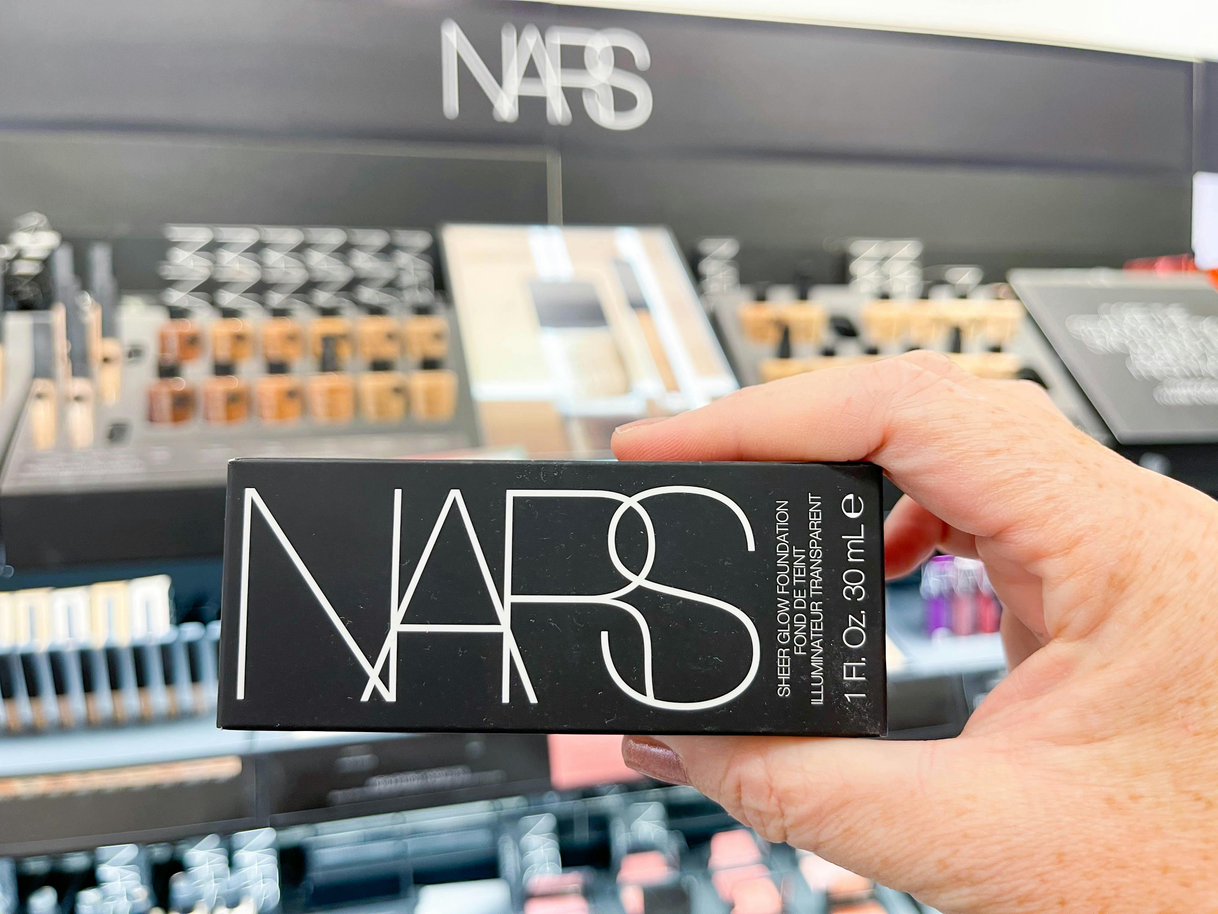 A person holding a box of nars foundation in front of the display in a Sephora store.