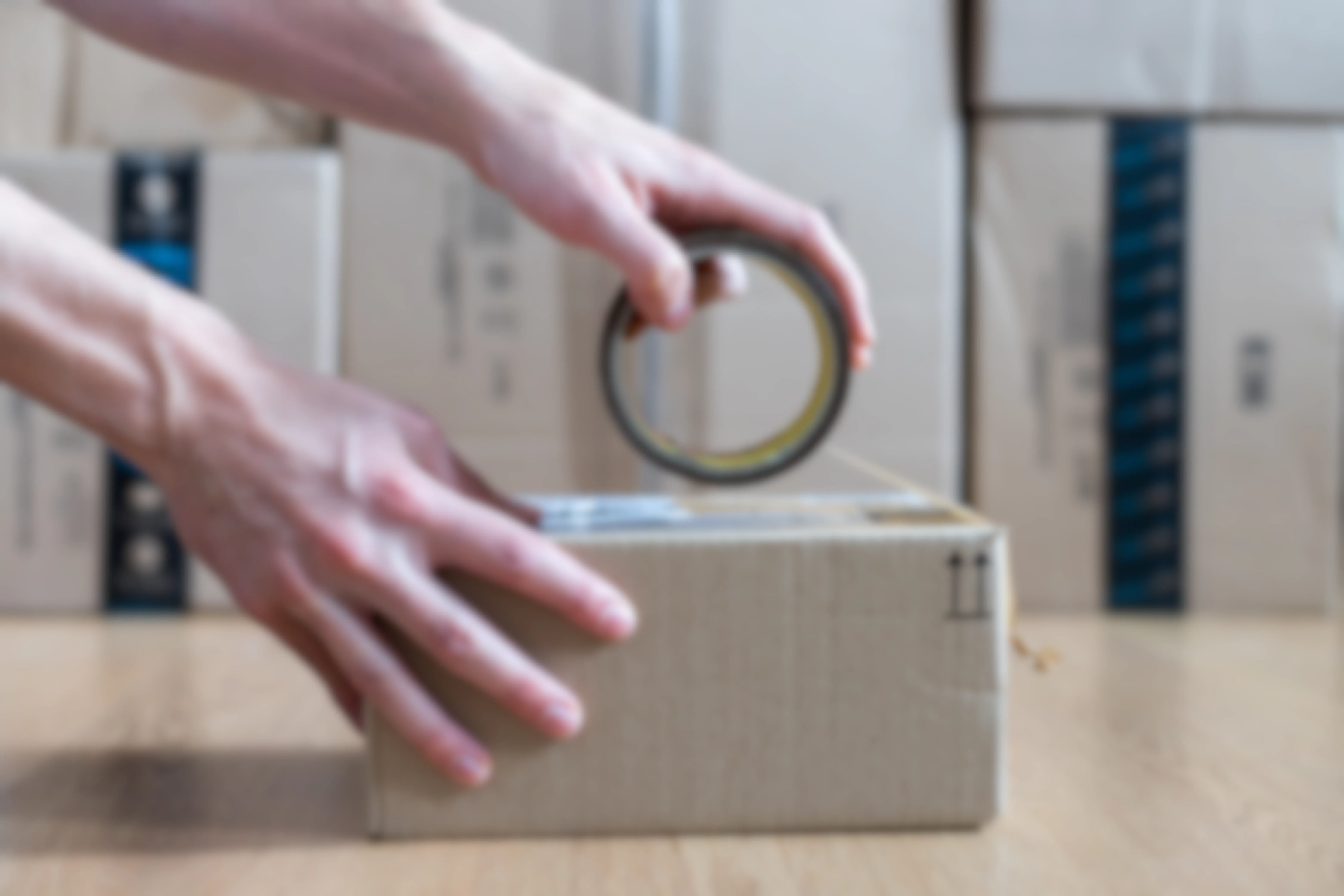 A man using packing tape to close a box. Cardboard boxes are stacked in the background.