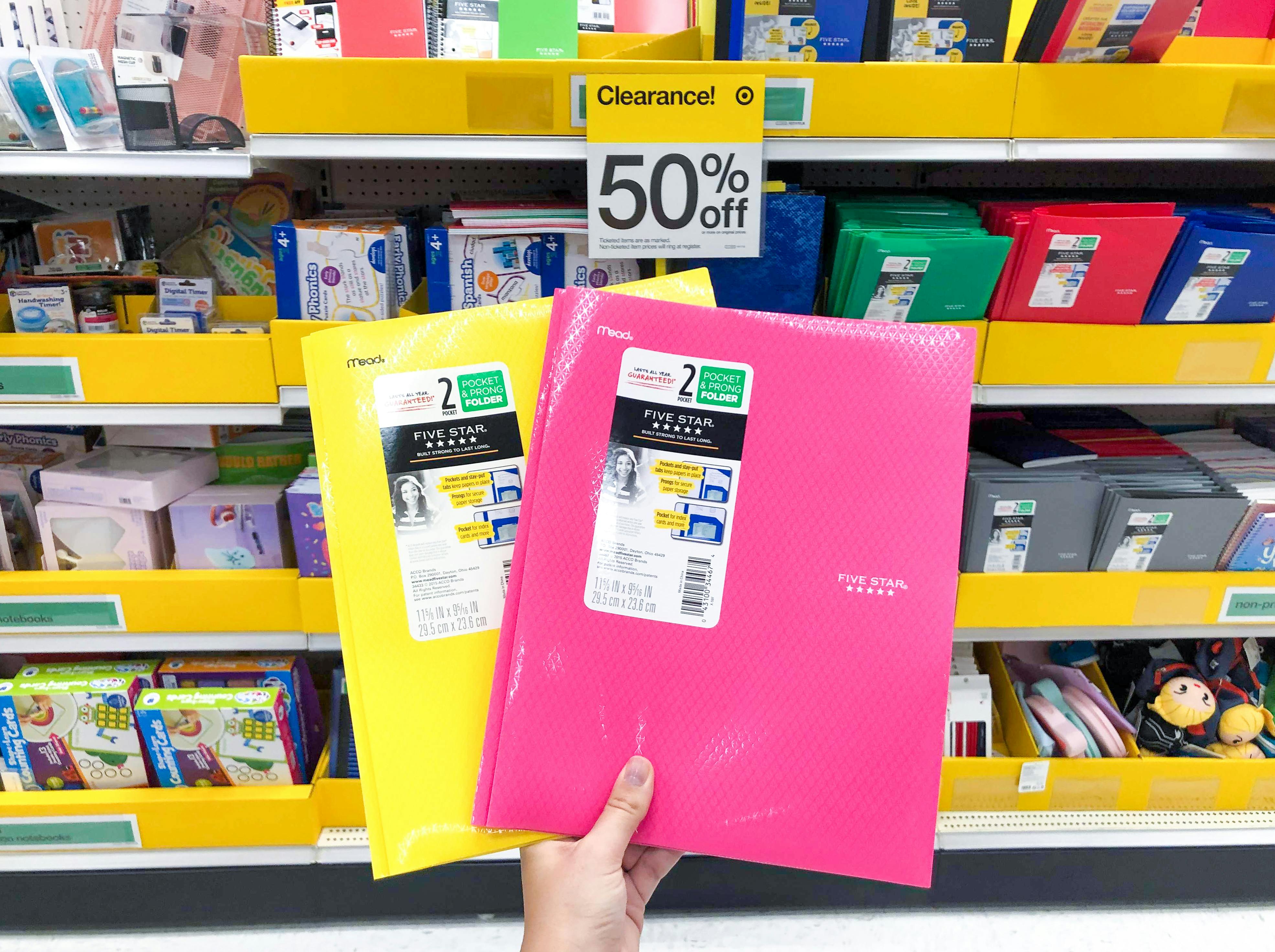 A person holding two folders in front of the back to school clearance section at Target. A sign that says "Clearance 50% off" is attached to the shelf in the background.
