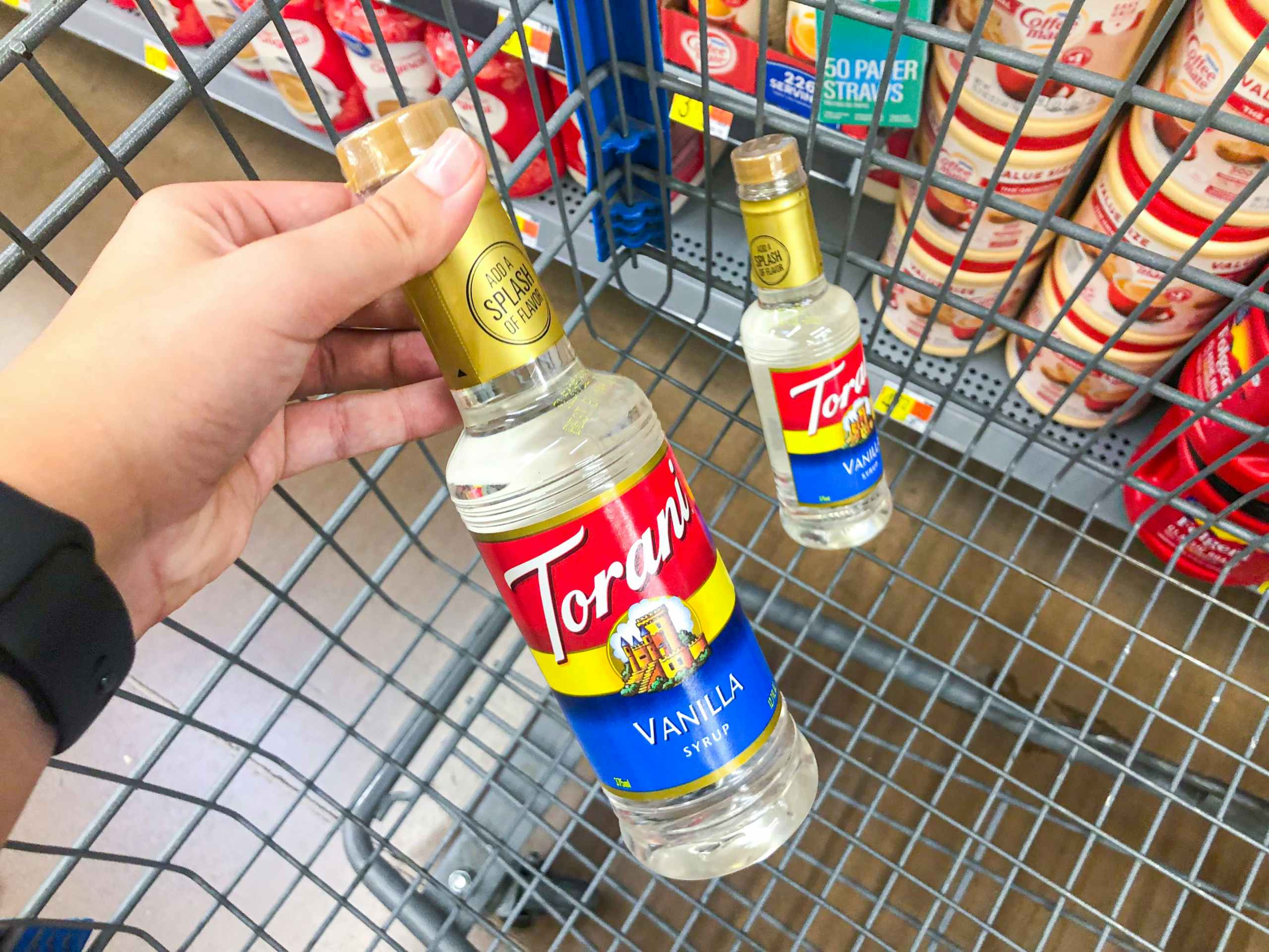 hand holding Torani bottle in front of another Torani bottle in a Walmart shopping cart