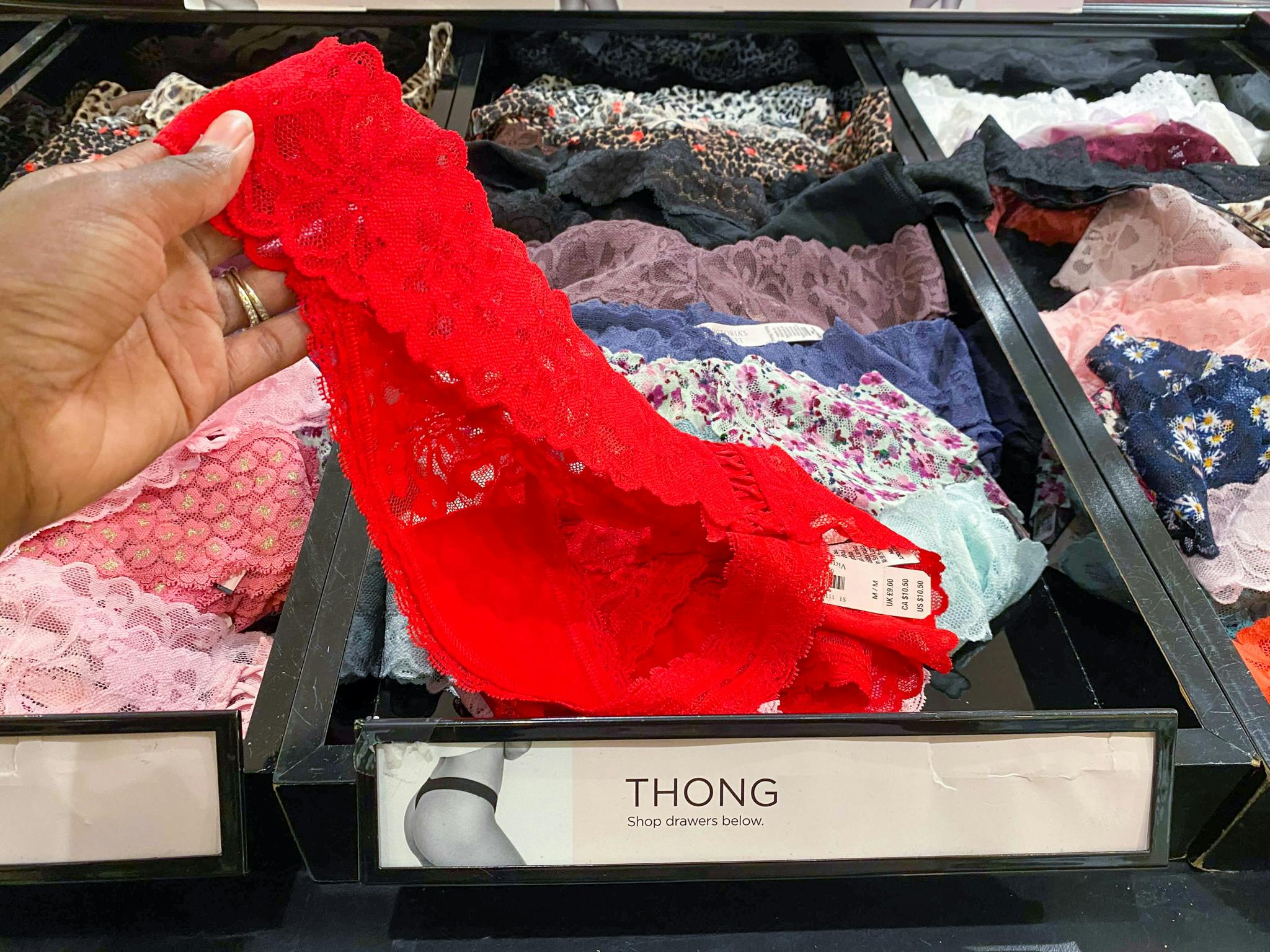 A person's hand picking up a lacy pair of underwear from a display at Victoria's Secret.