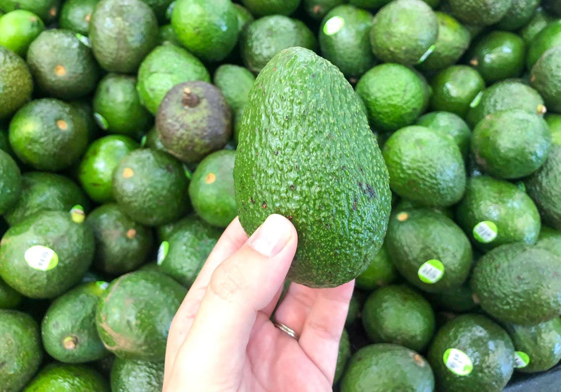 A person's hand holding an avocado above a pile of avocados.