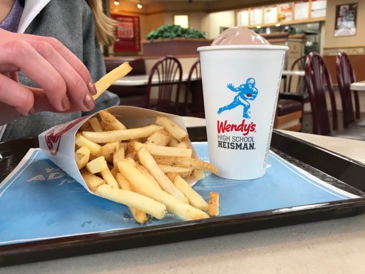 A person's hand picking up a french fry from a small box of Wendy's fries sitting on a tray next to a small chocolate frosty at Wendy's.