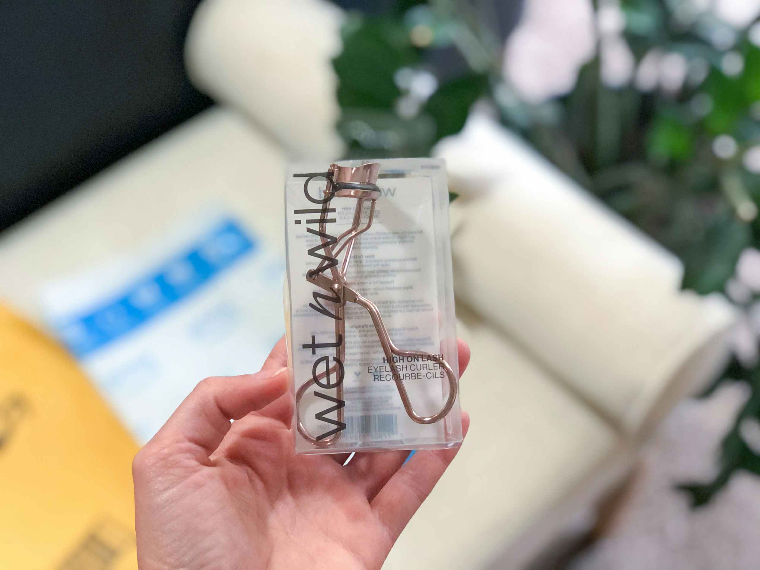 A hand holding a Wet n Wild eyelash curler in front of a delivery package.