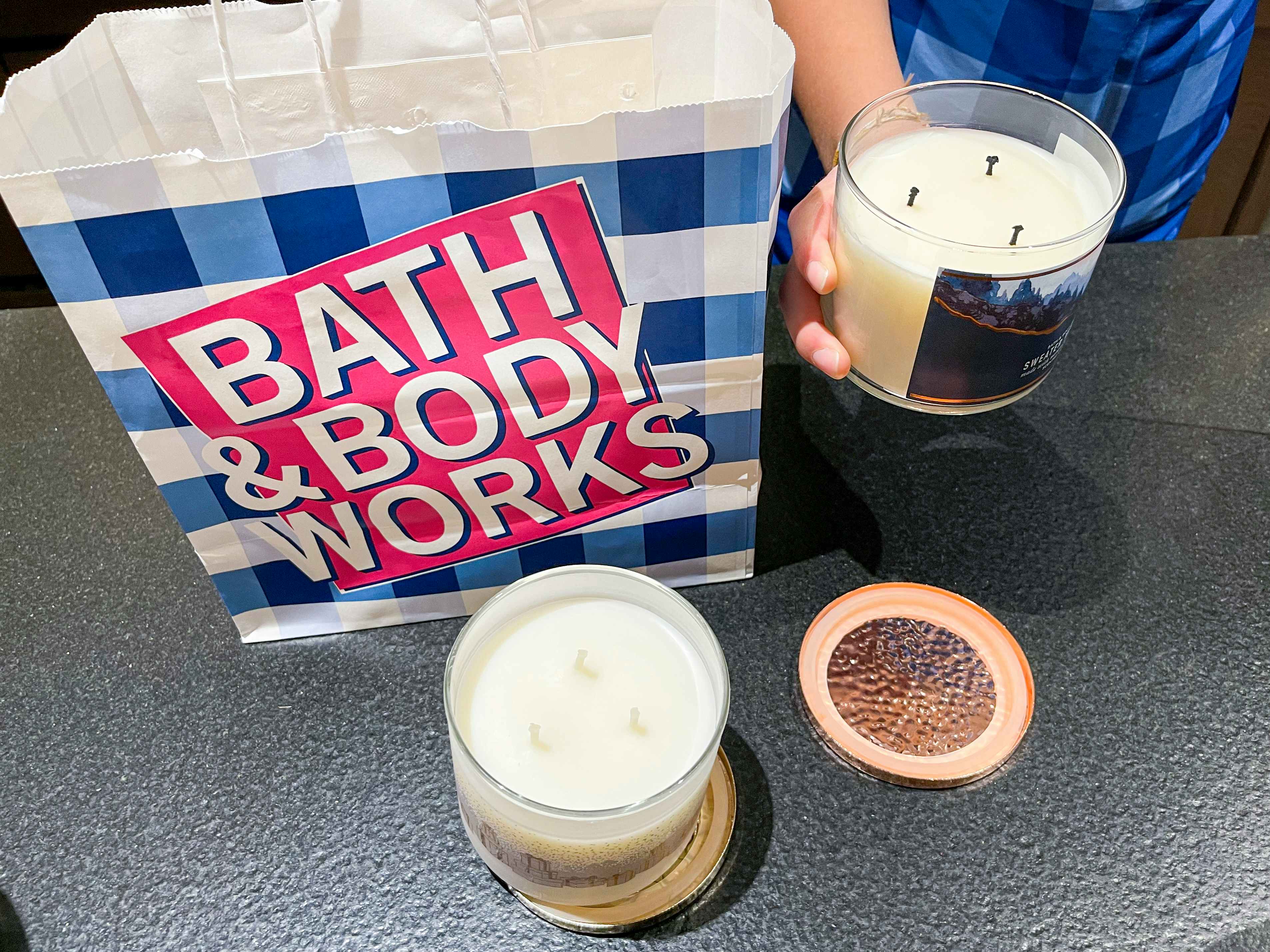 A person holding a 3-wick candle that has been lit before next to a Bath & Body Works bag and an unused 3-wick candle.