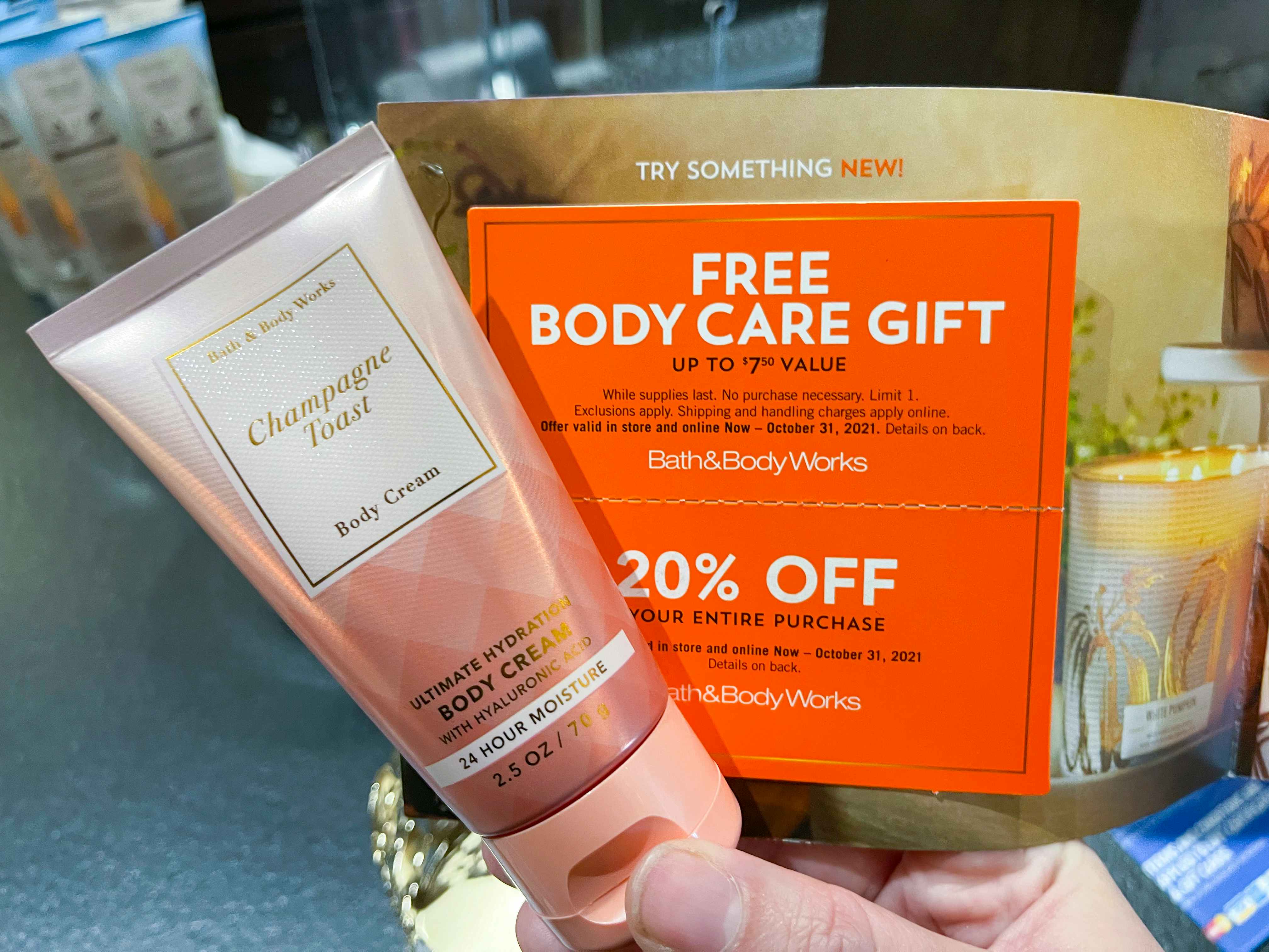 A person's hand holding a bottle of Bath and Body Works Champagne Toast body cream and a coupon for a Free Body Care Gift at Bath and Body Works.