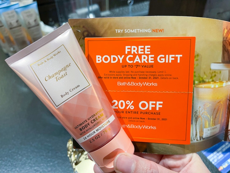 A person's hand holding a bottle of Bath and Body Works Champagne Toast body cream and a coupon for a Free Body Care Gift at Bath and Body Works.