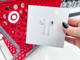A person's hand holding up a box of Airpods in front of a Target shopping cart.