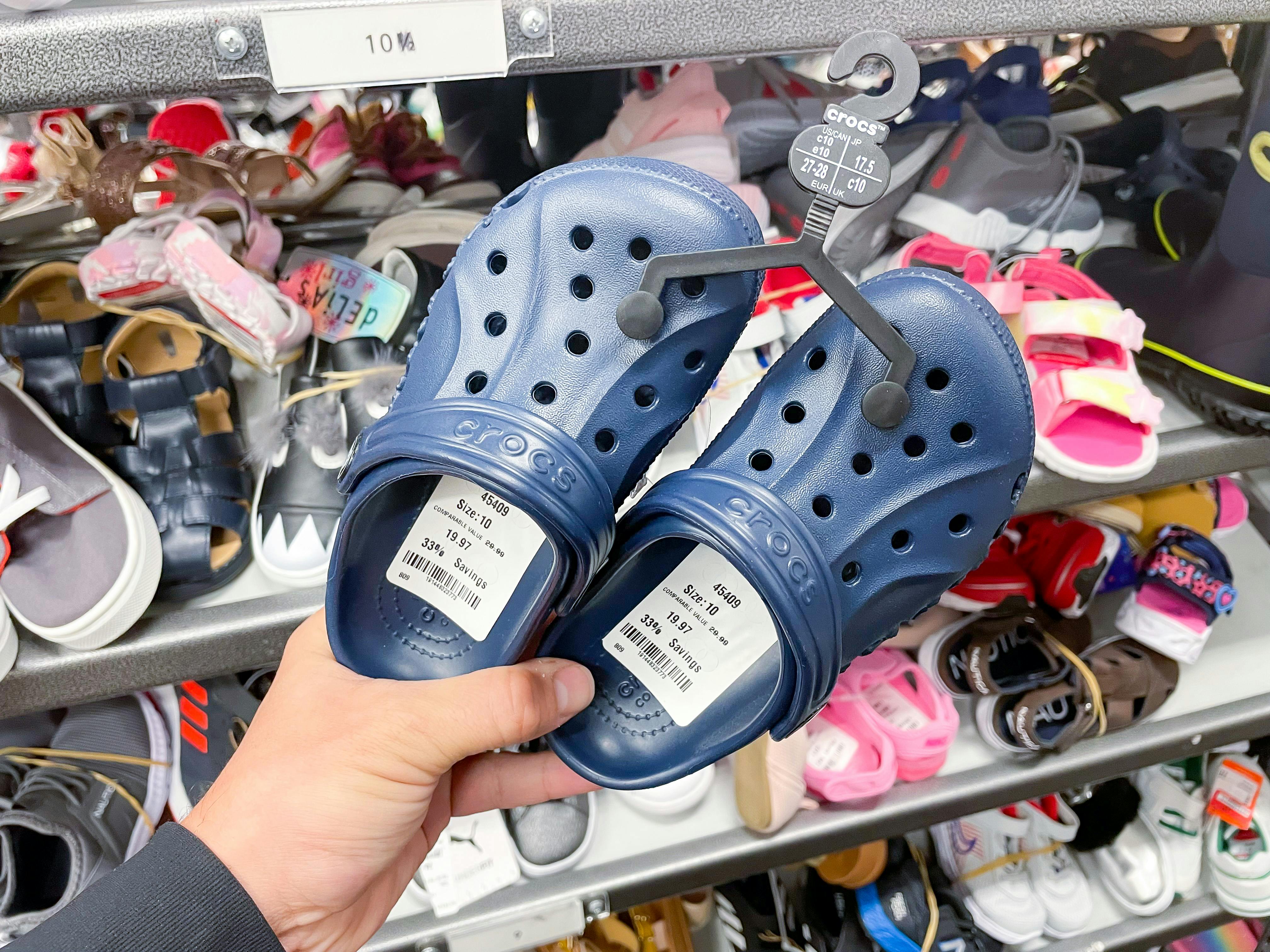 A person's hand holding a pair of kids Crocs in front of more kids shoes on a shelf.
