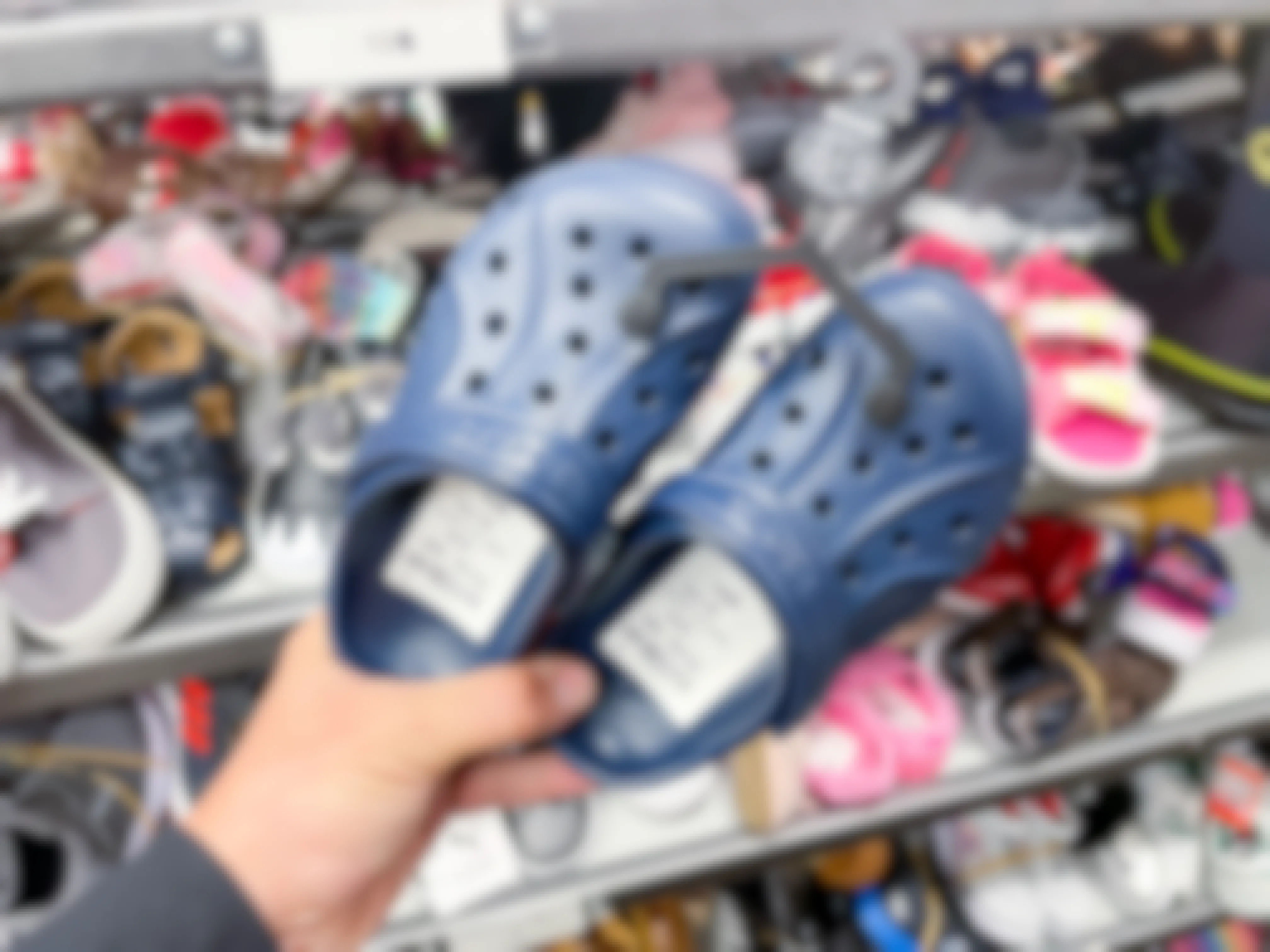 A person's hand holding a pair of kids Crocs in front of more kids shoes on a shelf.