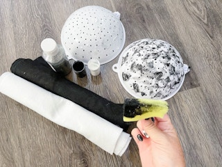 A person's hand holding a sponge with black paint on it in front of some materials for a DIY salt and pepper Halloween costume.