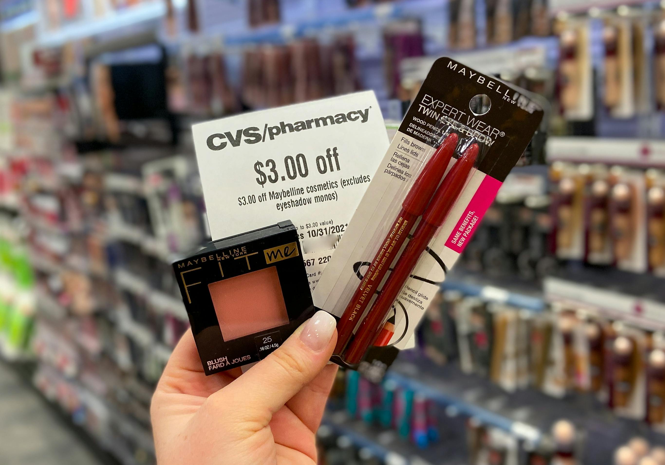 A person's hand holding up Maybelline makeup products with a coupon for $3 off at CVS.