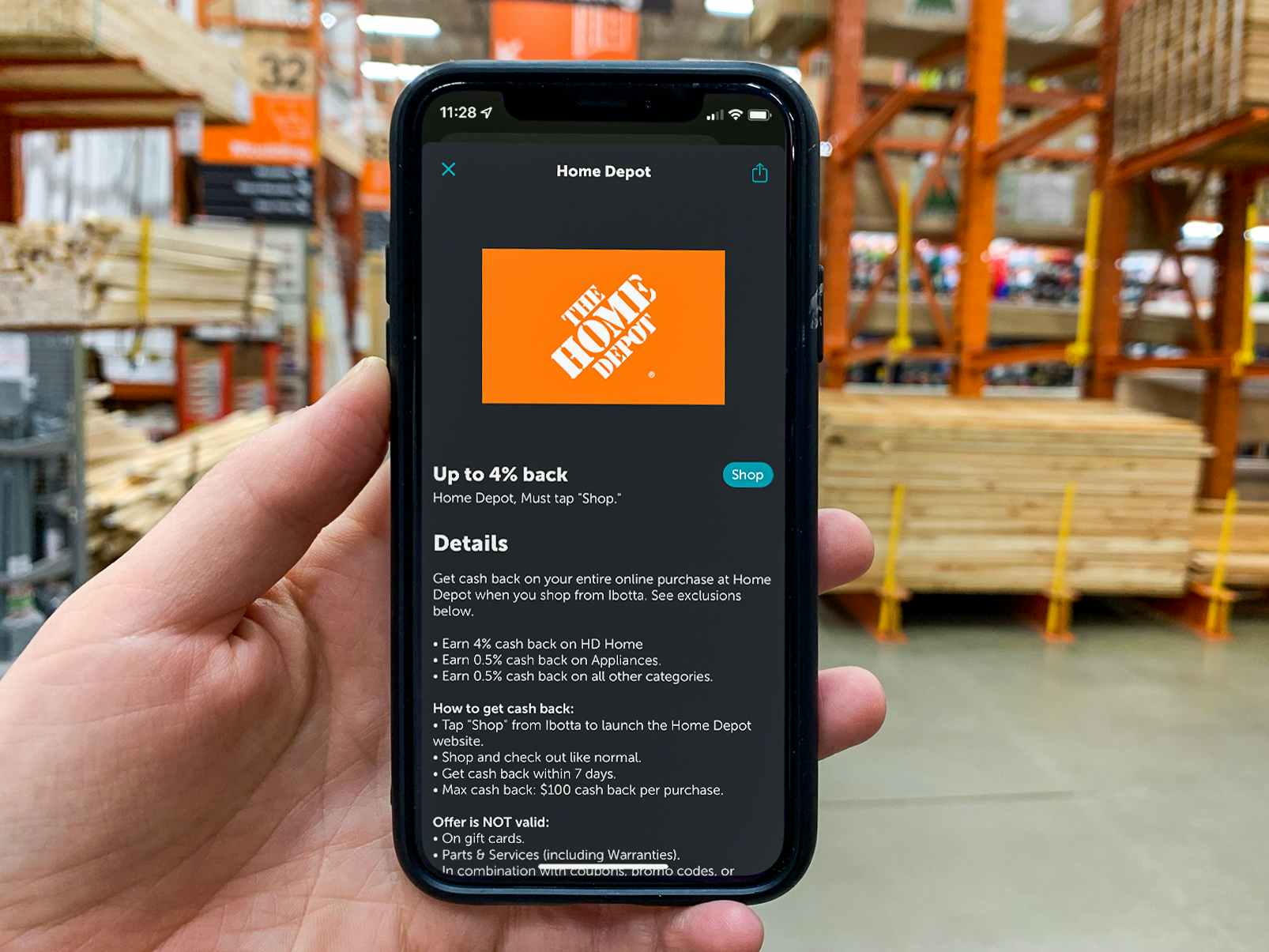 A person's hand holding an iPhone displaying the Home Depot offer page page on the Ibotta app in an aisle at Home Depot.
