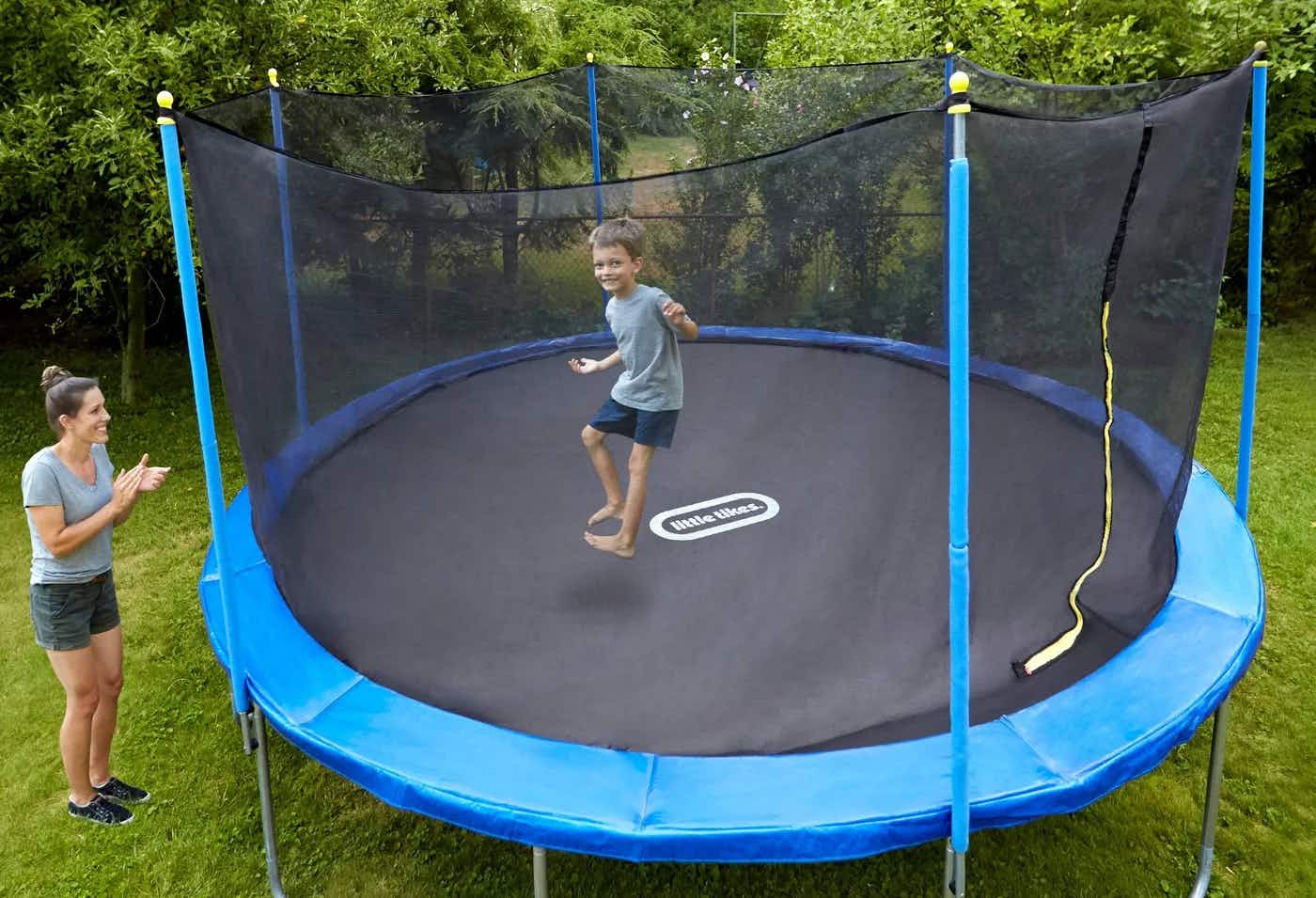 A child jumping on a Little Tikes brand trampoline in the backyard while the mom watches and smiles.