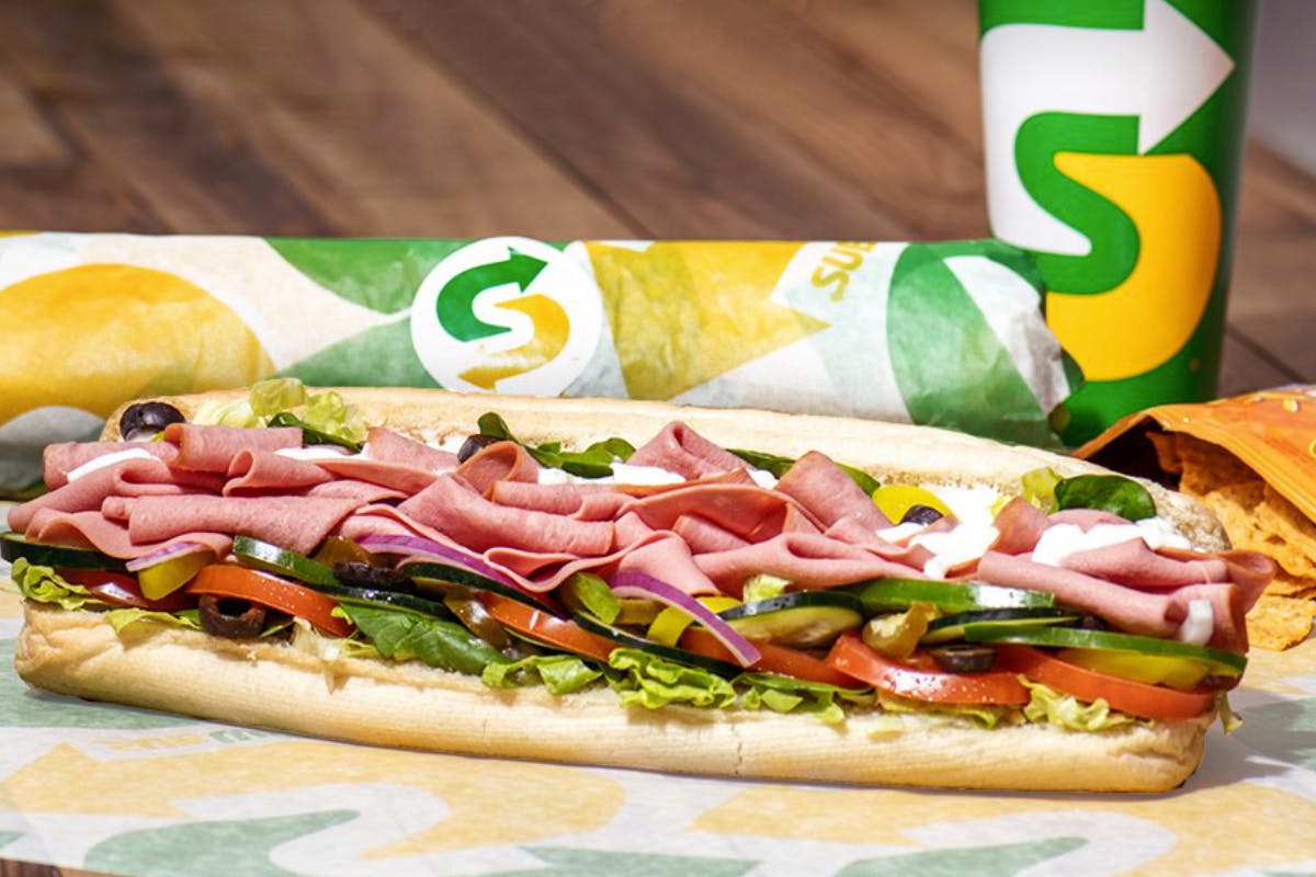 A Subway sandwich on a table with another wrapped sub and a drink