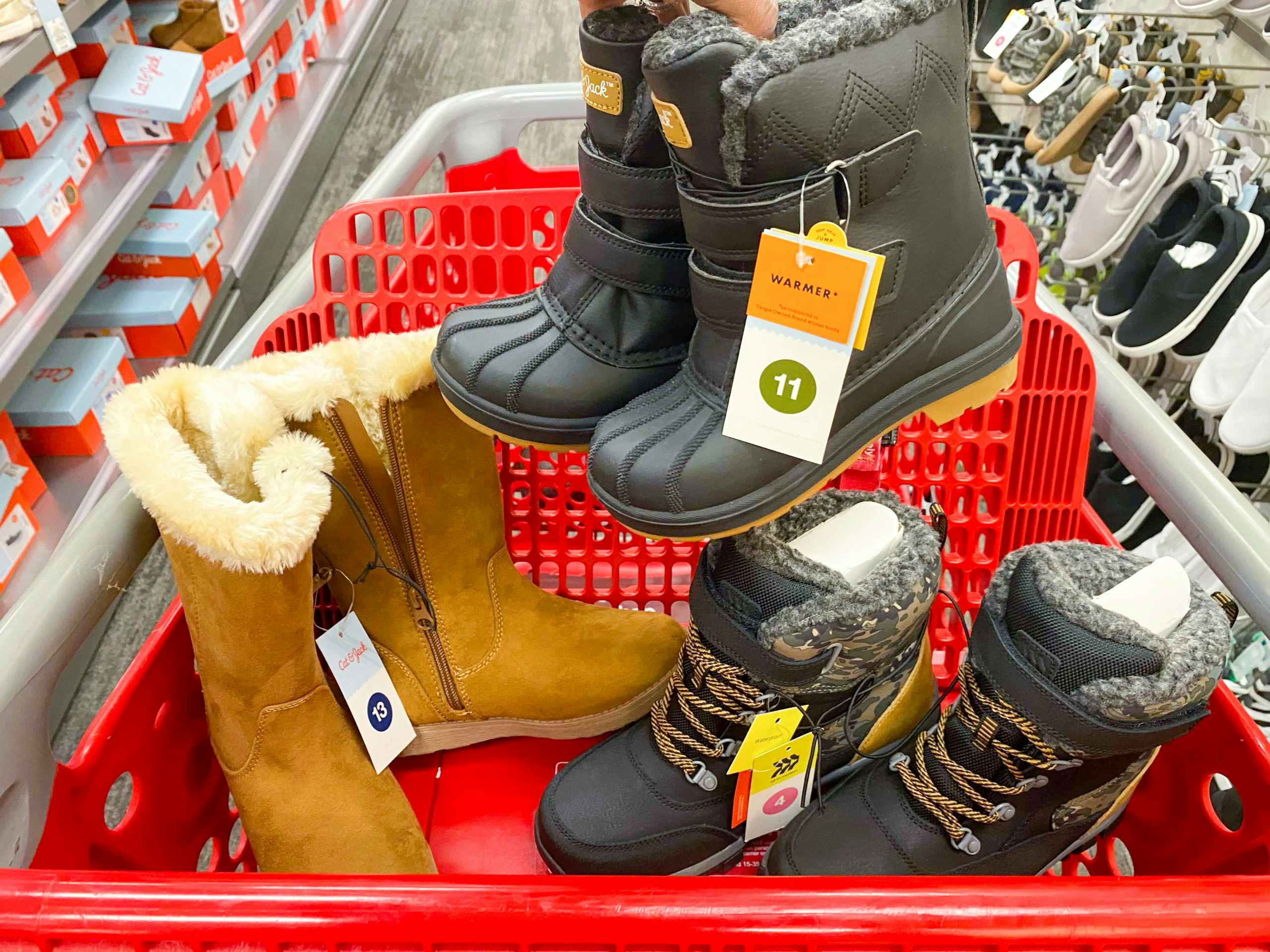 A person's hand holding a pair of children's boots above a Target shopping cart that has more children's boots in the basket, parked in the shoe aisle at Target.